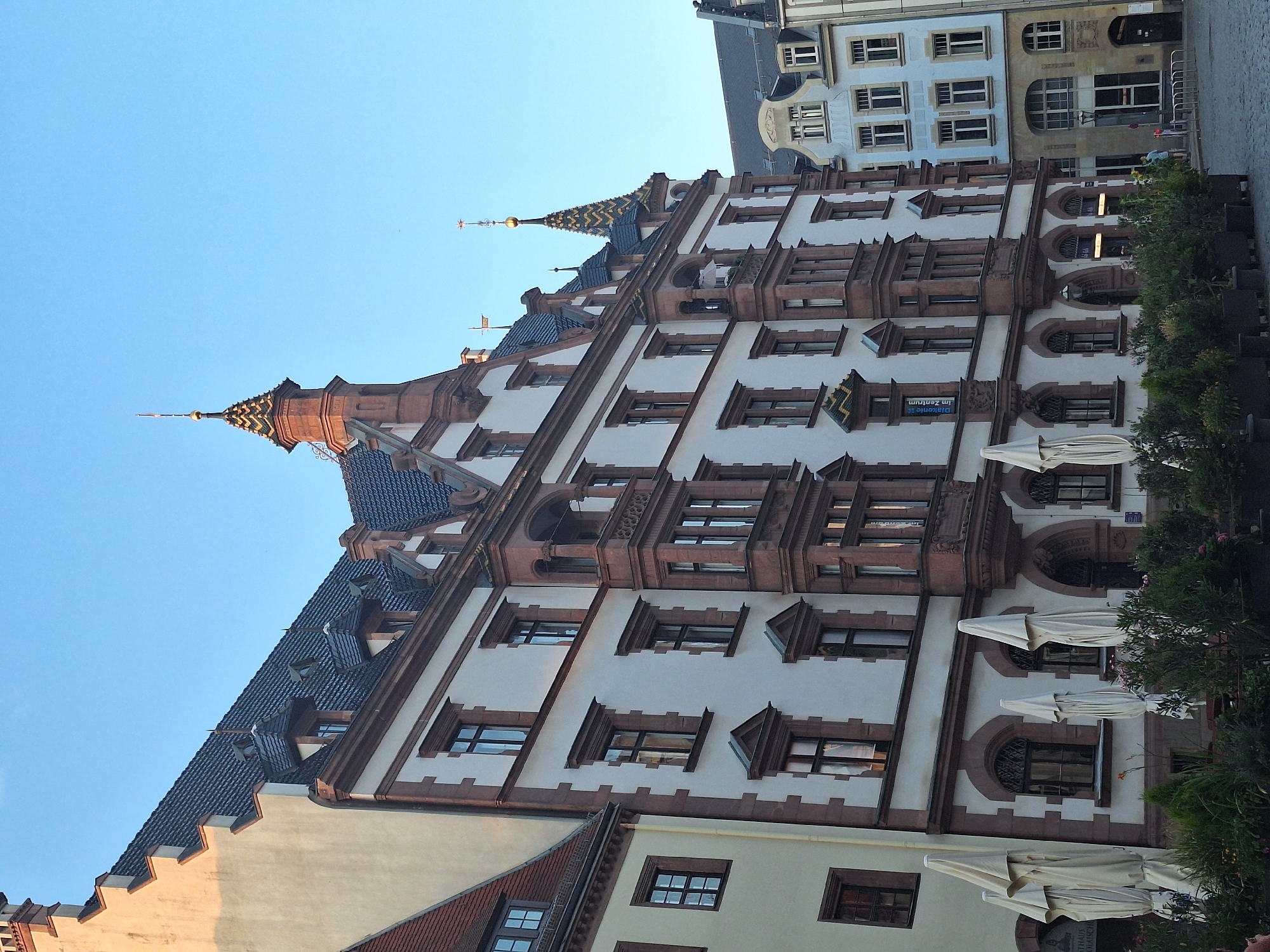 The image depicts the market square in Leipzig, Germany, on a clear day with a pale blue sky. The central focus is the historic Rathaus (City Hall) building, an impressive example of Renaissance architecture. Its white facade is adorned with reddish-brown sandstone accents around the windows and along the roofline. The building features multiple stories with numerous windows, some of which are arched. A prominent tower with a distinctive tiled spire, topped with a golden finial and star, rises from the building's structure. Dormer windows are visible on the steeply pitched, dark-tiled roof.
To the left of the Rathaus, a part of another building is visible, characterized by a pale yellow facade and a red-tiled roof with a gabled section. In front of the Rathaus, several white, closed umbrellas are arranged, suggesting an outdoor seating area for a café or restaurant. Lush green plants in planters line the foreground.
To the right of the Rathaus, a lighter-colored building with a pale blue facade is visible, also featuring multiple windows and a multi-story design. The ground level of this building has a more modern appearance with large windows and glass doors.
The ground is paved with cobblestones, typical of European city centers. In the background, to the right, a person in a light blue shirt is walking away from the camera on the cobblestone square. The scene is bathed in the warm light of either early morning or late afternoon, casting subtle shadows.