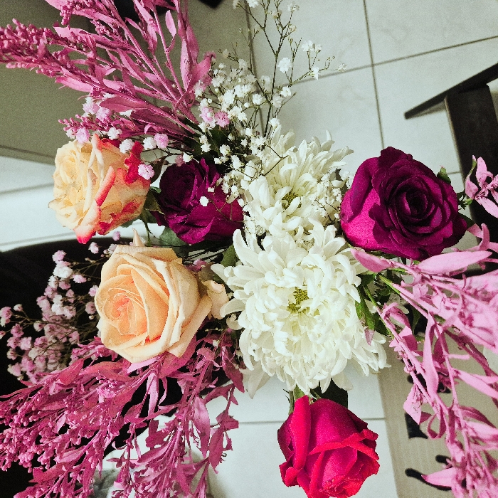 The scene depicts a vibrant floral bouquet presented in an indoor setting in Strasbourg, France. The bouquet comprises multiple flower types and colors. Dominant elements include five roses: two light peach-colored, one with prominent red-tipped petals; two dark magenta-hued; and one bright fuchsia-pink. Centrally located are several large, white chrysanthemums with visible green centers. Numerous small, white and light pink baby's breath flowers are interspersed throughout the arrangement, along with distinct branches of a feathery plant dyed or painted a bright fuchsia-pink, extending outwards. The bouquet is viewed from a slightly elevated perspective. The immediate background on the right reveals a light-toned, tiled floor with visible grout lines. A dark, out-of-focus object, possibly a chair back, is partially visible in the lower right corner. The upper left and top background areas are dark and indistinct. No people are present, and no actions or interactions are depicted.