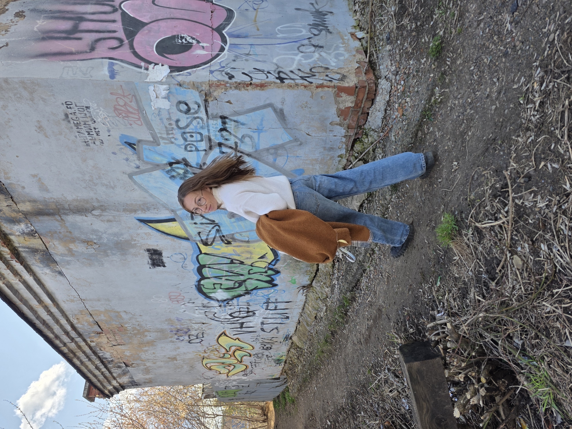 The image, rotated 90 degrees clockwise, shows a young woman posing outdoors against a heavily graffitied wall, likely in Chaikovsky, Russia.
The subject is a young woman with long, light brown hair that appears to be in motion, suggesting a dynamic pose or a slight breeze. She wears a white turtleneck sweater, light blue wide-leg jeans, and round spectacles. In her left hand, she holds a crumpled brown garment, possibly a jacket or a bag. Her expression is neutral as she looks towards the camera.
The scene is an urban or suburban environment with a raw, artistic, and somewhat dilapidated character. The primary background is a textured concrete wall, which is severely cracked and has large patches of peeling plaster, revealing exposed brickwork underneath. This wall is extensively covered in various layers of colorful graffiti and black tags. The ground in front of the wall is unpaved, composed of dirt, small stones, and sparse, dry vegetation including twigs and weeds. In the distance, above the structure, a clear blue sky with some white clouds is visible, along with bare tree branches, suggesting a spring or late autumn setting. A dark wooden post stands upright in the foreground on the right.
The visible activity is a posed photograph. The time of day is daylight, indicated by the bright sky and natural lighting.
Visible text and notable graffiti elements on the wall include:
* A large pink and black stylized tag on the upper section.
* Black text that appears to read "HERAMEGAFO" (or a variation) on the left side.
* A blue tag that includes "Pase".
* A green and yellow stylized tag near the center.
* Another yellow and green tag below which "WHO STUFF" is partially legible.
* Numerous other illegible scribbles and abstract graffiti.