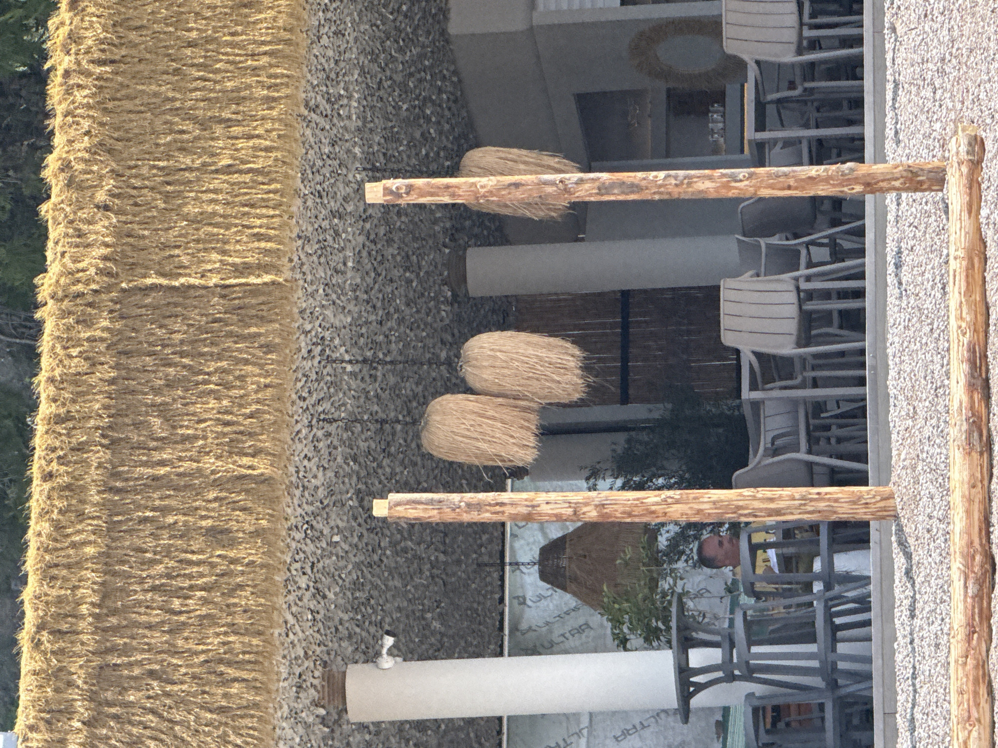 The image shows an outdoor seating area, potentially part of a restaurant or bar, located in Budva, Montenegro. The area is characterized by its rustic design, featuring a thatched roof and wooden support beams. The flooring appears to be made of small stones or pebbles. Several light-colored chairs are arranged around tables, suggesting a relaxed atmosphere. In the background, glimpses of greenery indicate the presence of vegetation. A man is partially visible seated at one of the tables. Decorative straw lamps and a straw wreath add to the ambiance. The structural elements combined with the seating suggest a commercial establishment catering to tourists or locals.