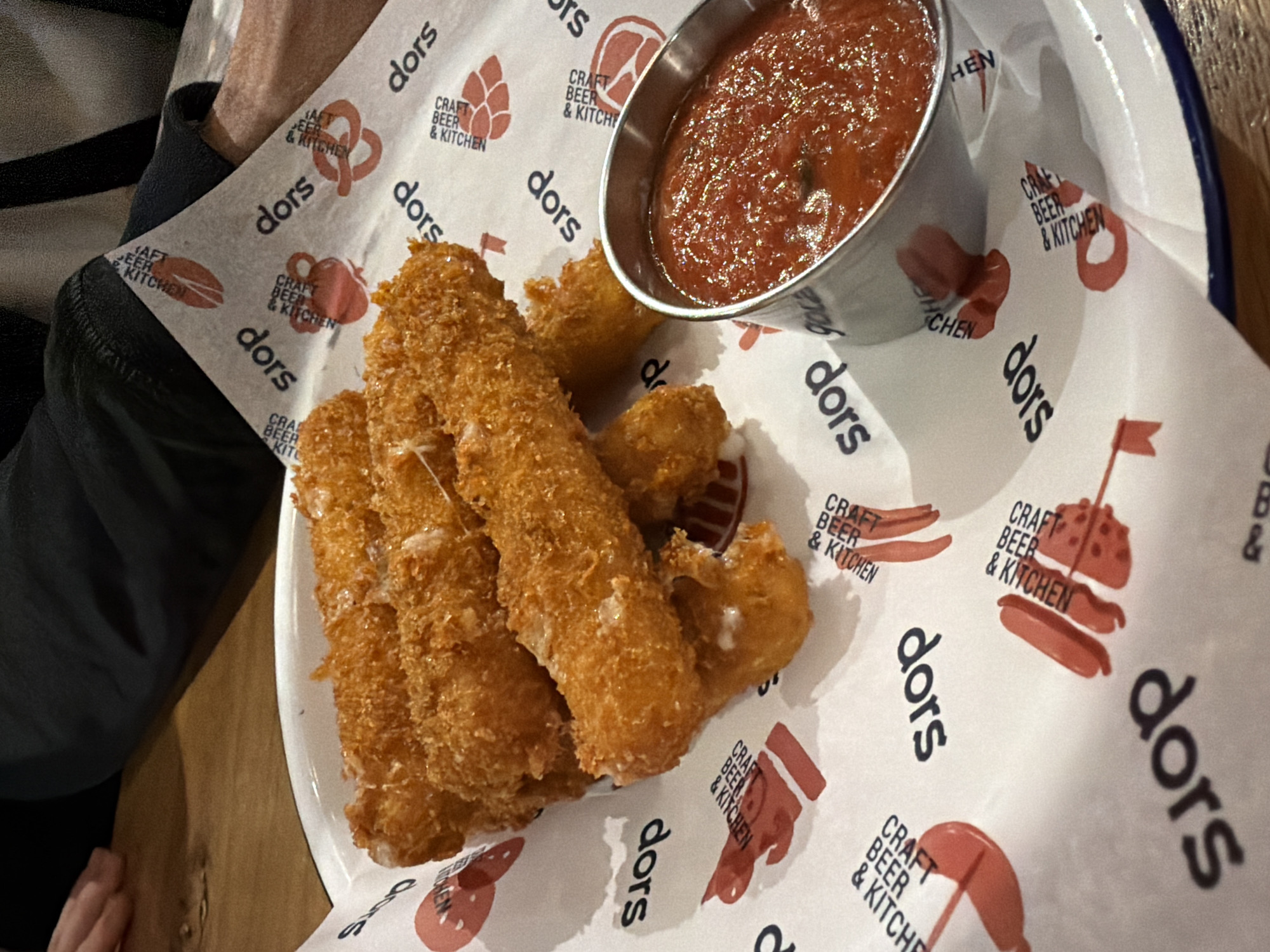 The image shows a plate of fried mozzarella sticks with a small metal cup of red tomato sauce on patterned paper. The paper has the text "dors CRAFT BEER & KITCHEN" printed on it, along with illustrations of beer-related items. A person's arm and hand are visible on the left side of the frame. The food appears to be served at a location called "dors Craft Beer & Kitchen", which is potentially located in Yerevan, Armenia.