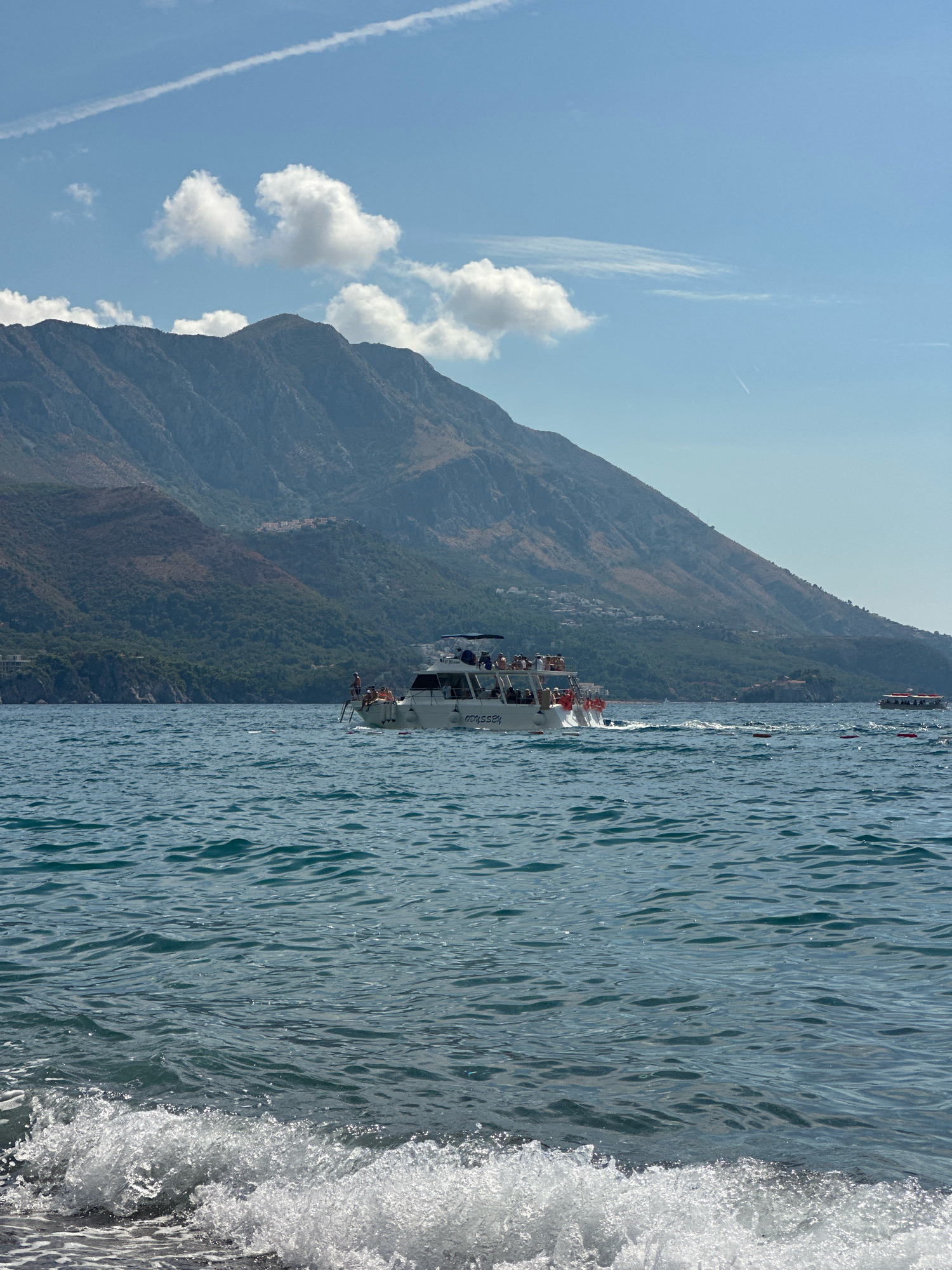 In the city of Boreti, Montenegro, on a clear, sunny day, a white tourist boat with people on deck cruises across the blue sea. The boat, named "ODYSSEY," is positioned in the middle ground of the image. In the background, a large, verdant mountain slopes down to the coastline, dotted with buildings. The foreground features a small wave lapping at the shore. The sky above is a bright blue, with a few white clouds and a faint contrail from an aircraft. Another smaller boat is visible in the distance to the right.