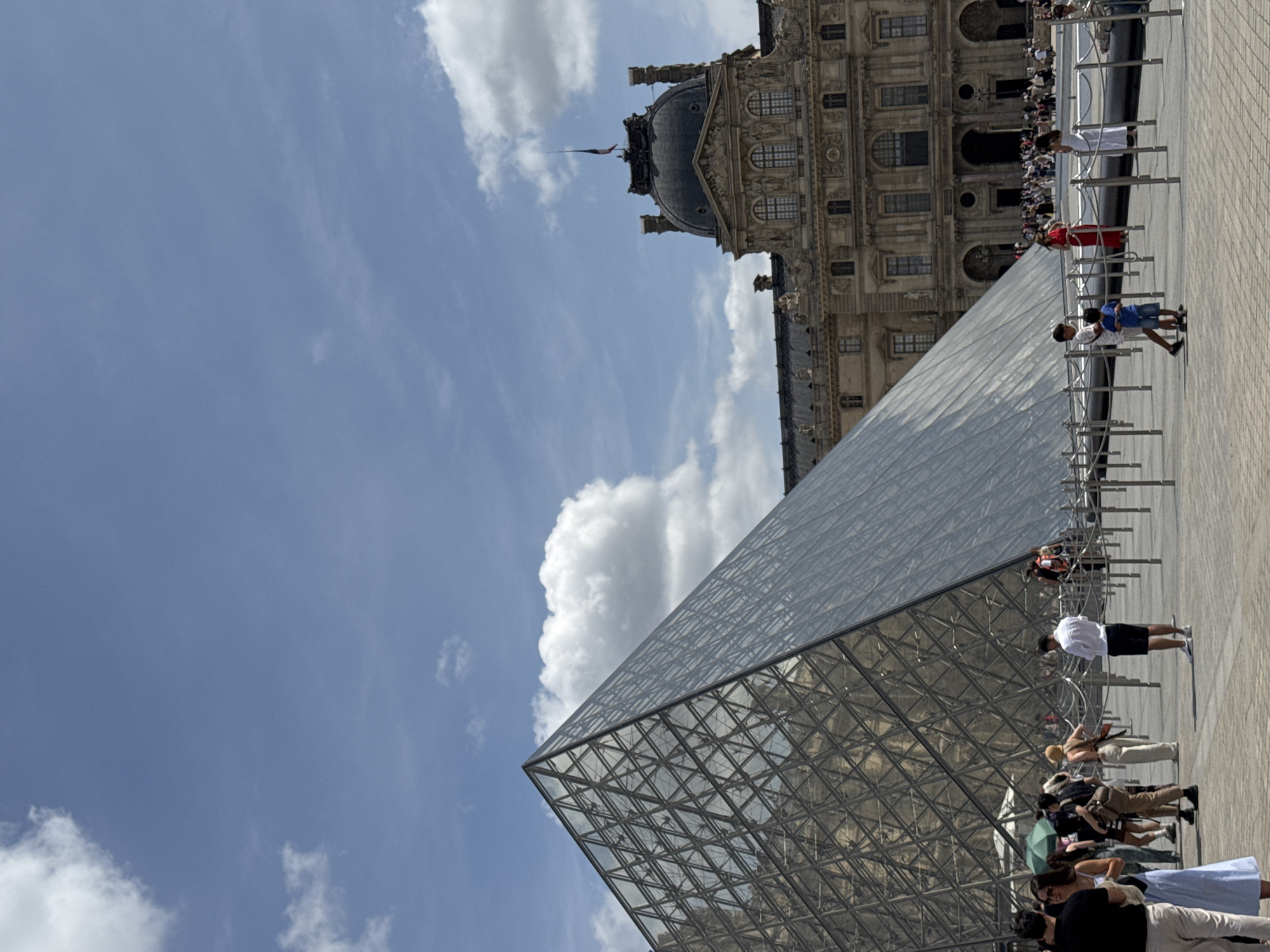 The image depicts the iconic glass pyramid entrance to the Louvre Museum in Paris, France. The pyramid, a modern architectural addition to the historic palace, stands prominently in the foreground, its geometric structure reflecting the surrounding sky and building. Behind the pyramid, the grand facade of the Louvre Palace, with its ornate details and classical architecture, is visible.
The scene is bustling with activity, characteristic of a popular tourist attraction. A crowd of people, dressed in casual attire, are gathered in the plaza. Some are walking, others are standing in small groups, and a few are posing for photographs. Several individuals are seen interacting with each other, possibly discussing directions or sharing their experiences.
The environment is an open, paved plaza, indicative of a public space designed for pedestrian access and gathering. Ropes and stanchions are set up, suggesting a queuing system or a designated pathway for visitors. The sky above is a bright blue, with scattered white clouds, suggesting fair weather. The overall impression is one of a lively and historic urban setting, with the Louvre Museum serving as the central point of interest.