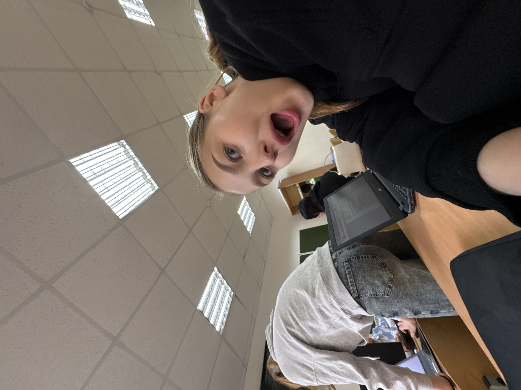 The image depicts an indoor scene, likely a classroom or study area, judging by the presence of ceiling lights, desks, and laptops. A young woman with blonde hair, wearing a black sweatshirt, is positioned in the upper part of the frame with an open mouth and is looking directly at the camera. Another person wearing a white long sleeved t-shirt and jeans is located in the bottom center and is on their laptop. There is another person wearing a black sweatshirt behind the person with the white long sleeved shirt. Laptops are positioned on the wooden desktops. The location is speculated to be the city of Chaikovsky, Russia. The image seems to have been taken from an unusual perspective, possibly with the camera held above the subjects.