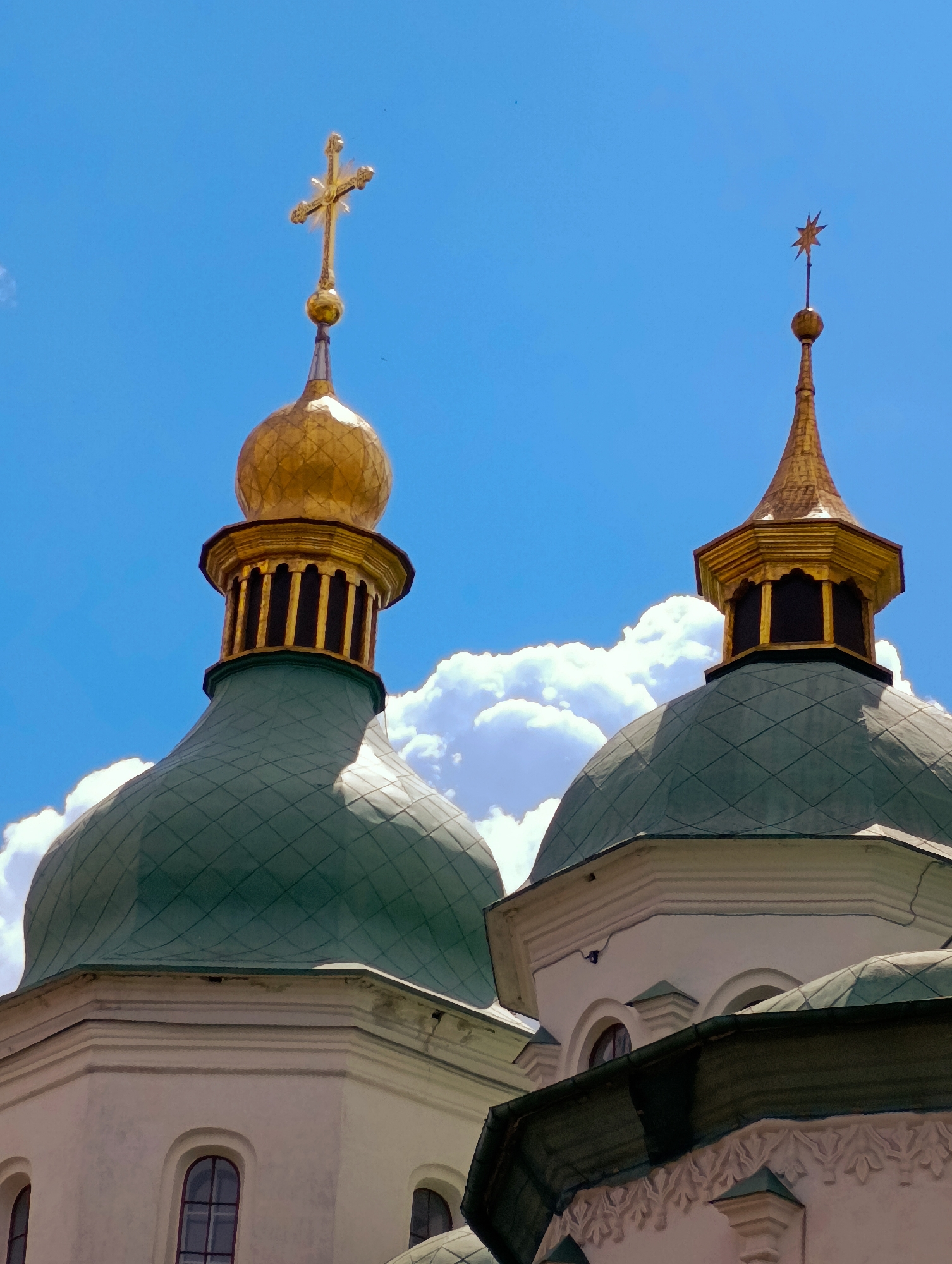 The image shows the golden domes and green onion domes of St. Sophia's Cathedral, a historic Orthodox Christian cathedral located in Kyiv, Ukraine. The sky is a vibrant blue with fluffy white clouds. The domes are ornate, with intricate patterns and crosses topping them. The architecture is characteristic of Eastern Orthodox churches, with a blend of Byzantine and Ukrainian Baroque styles. The surrounding environment is not visible in the frame, but the focus on the cathedral's impressive structures suggests a grand and significant religious site. The image captures a clear, sunny day, highlighting the architectural beauty of the cathedral.