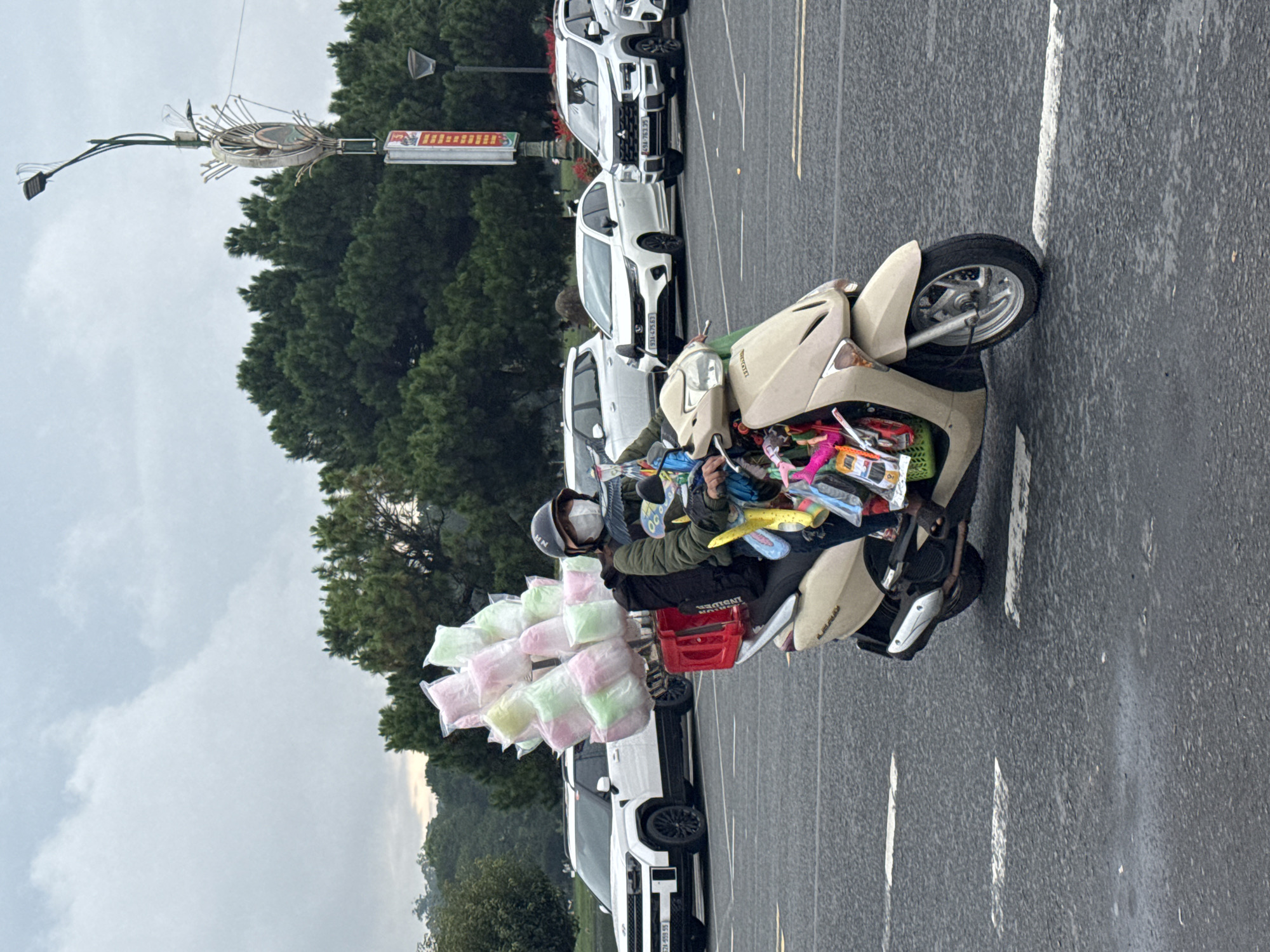 A street vendor on a beige Honda scooter drives along a paved road in Da Lat, Vietnam, under an overcast sky. The vendor is wearing a dark jacket, a helmet, and a white face mask, focusing on the road. The scooter is heavily loaded with goods for sale; a large stack of individually packaged cotton candy bags in pastel pink, green, and white is attached to the rear, while colorful items, possibly toys or small souvenirs, are packed in baskets or bags in front of the seat.
On the right side of the road, several white cars are parked, including an SUV and sedans, with visible Vietnamese license plates such as "51G-763.99", "51A-475.63", and "51H-695.55". A dense line of green trees, likely pine trees, forms the background, with distant hazy mountains visible, characteristic of Da Lat's landscape. To the left, a street light pole features an ornate circular decoration and a red rectangular sign with white text, possibly reading "ĐẠI NAM", though it is difficult to discern clearly. The road has visible white parking lines and a yellow lane divider. The scene captures a typical moment of mobile street vending in Vietnam during daytime cloudy weather.