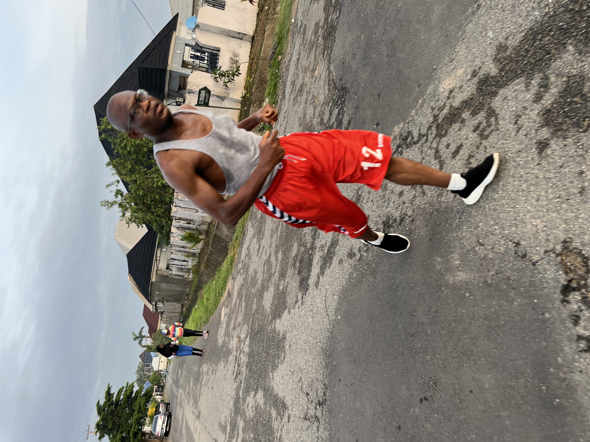 The image captures a group of people jogging on a street in Abuja, Nigeria. The primary subject is a man in the foreground, wearing a grey tank top and red shorts with the number "12" printed on them. He is in motion, appearing to be mid-stride. In the background, two other individuals are also jogging, with one wearing a hijab and the other a brightly colored hoodie. The street is paved and appears to be a residential area with houses and trees lining the sides. The sky is overcast, suggesting it may be early morning or late afternoon. A street sign reading "G.O. OBASOHAN STREET" is visible on the right side of the image, confirming the location as Abuja. The overall scene depicts an active, outdoor community scene.