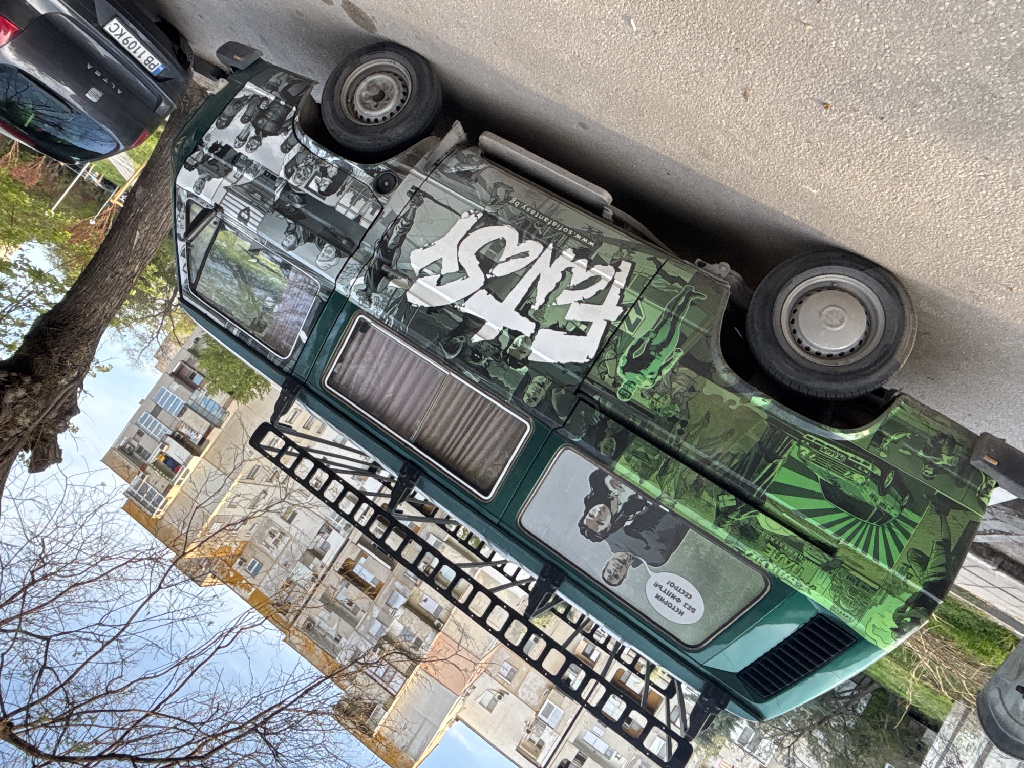 The image shows a green and black "Fantasy" branded van parked on a city street. The van has various graphic designs on it, including a white "Fantasy" logo, images of people, and comic book-style artwork. It also has a roof rack. In the background, there is a multi-story building, possibly residential, and a tree with bare branches. Another car, a dark grey hatchback, is parked adjacent to the van. The car's license plate is visible and reads "PB 1109 KC". The location is likely in Plovdiv, Bulgaria, due to the license plate and the branding for the Sofia Fantasy Film Fest.