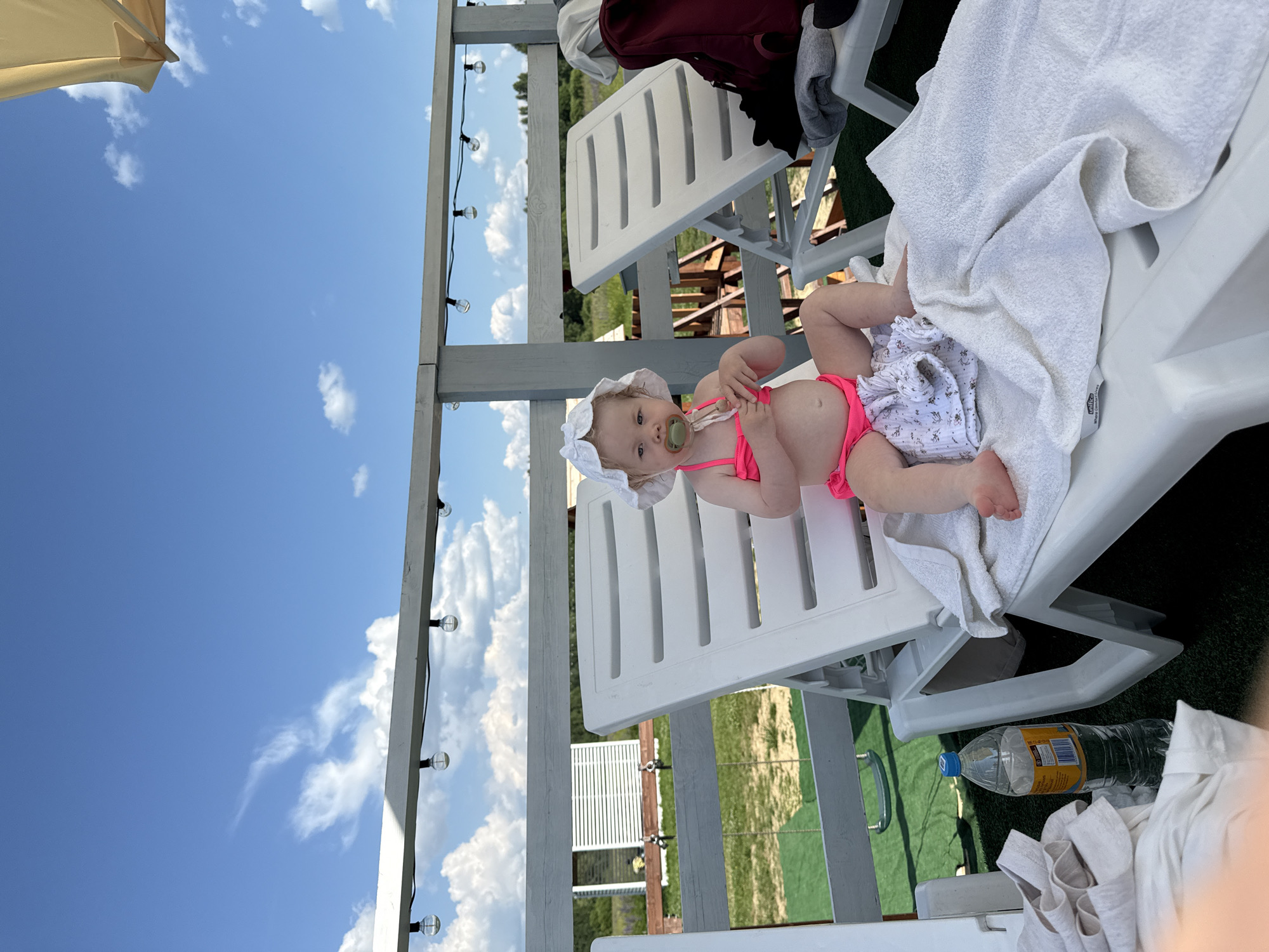An infant wearing a pink swimsuit and a white sun hat sits on a white lounge chair. A pacifier is in the infant's mouth. The infant is holding a small object in their hands. A white towel is draped over the lounge chair. In the background, there is a wooden fence, green grass, and a building with white siding. String lights are visible overhead. A plastic bottle of water is on the ground next to the lounge chair. The scene appears to be an outdoor recreational area, potentially near Колокша, Russia, given the reference. The sky is blue with scattered white clouds.