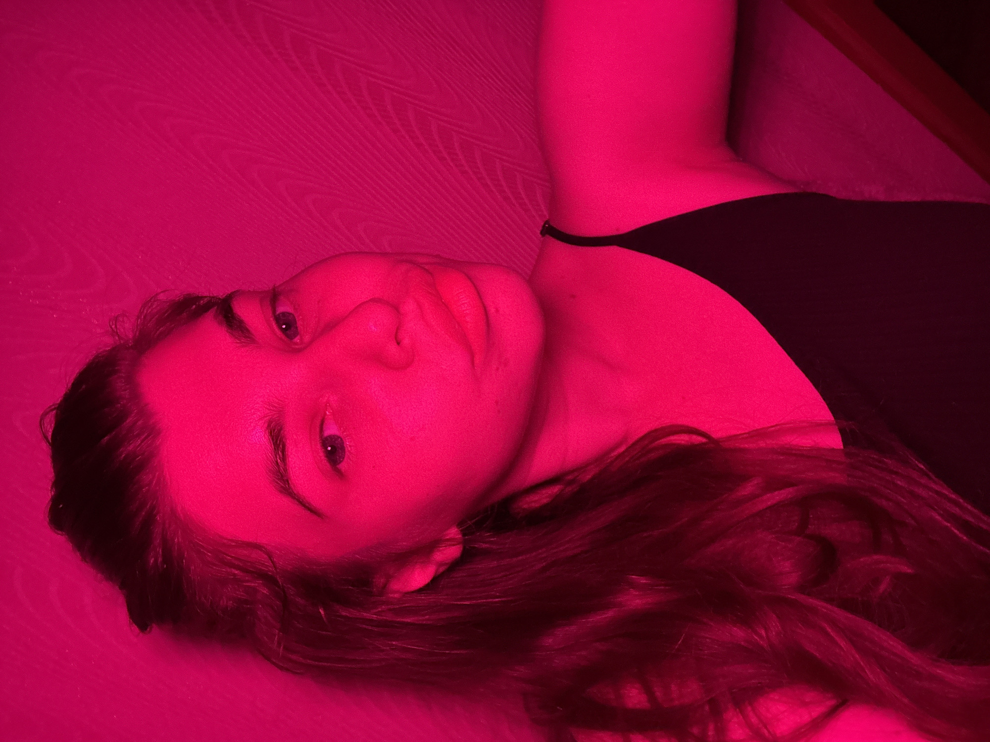 A young woman with dark hair pulled up is captured in a close-up, angled shot. She is wearing a black spaghetti strap top. The lighting in the scene is a vibrant, saturated pink, casting a strong hue over her face and the textured wall behind her. Her features are softened by the pink light, and her expression appears neutral. The image is taken indoors, with the textured wall suggesting an interior space. There is no indication of the city of Мещёры, Russia, in the provided image or its crops.