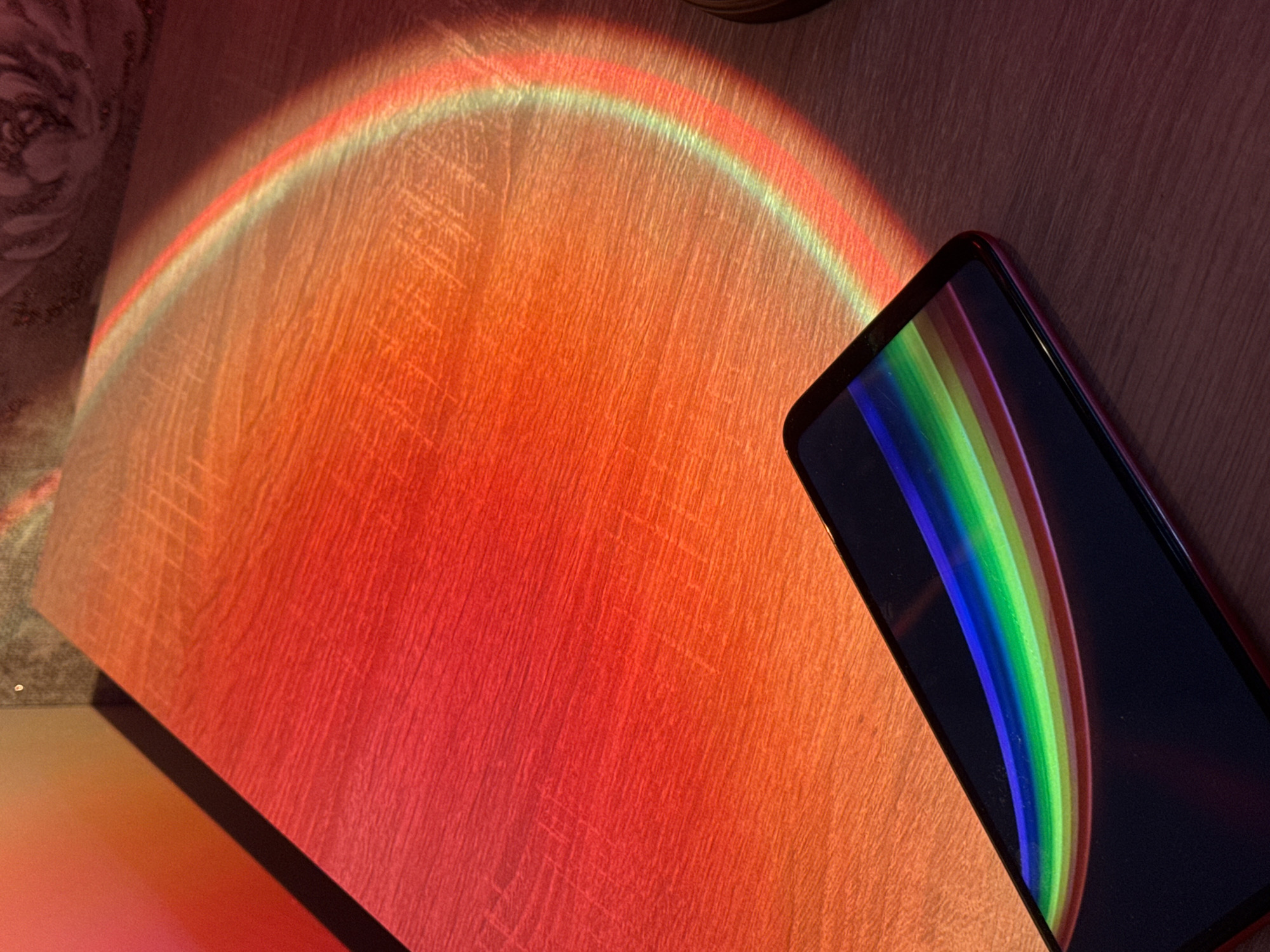The image shows a close-up, high-angle shot of a wooden surface with a sunset lamp projecting a rainbow arc. In the foreground, a black smartphone with a red border displays a rainbow reflection on its screen. The wooden surface exhibits a prominent grain pattern and is illuminated by the warm, vibrant colors of the projection, transitioning from deep red to orange and then to a faint green at the top of the arc. The overall scene is one of artificial light creating a colorful display on a textured surface, with the smartphone acting as a reflective element. There are no people or specific city landmarks visible, thus the location cannot be determined.