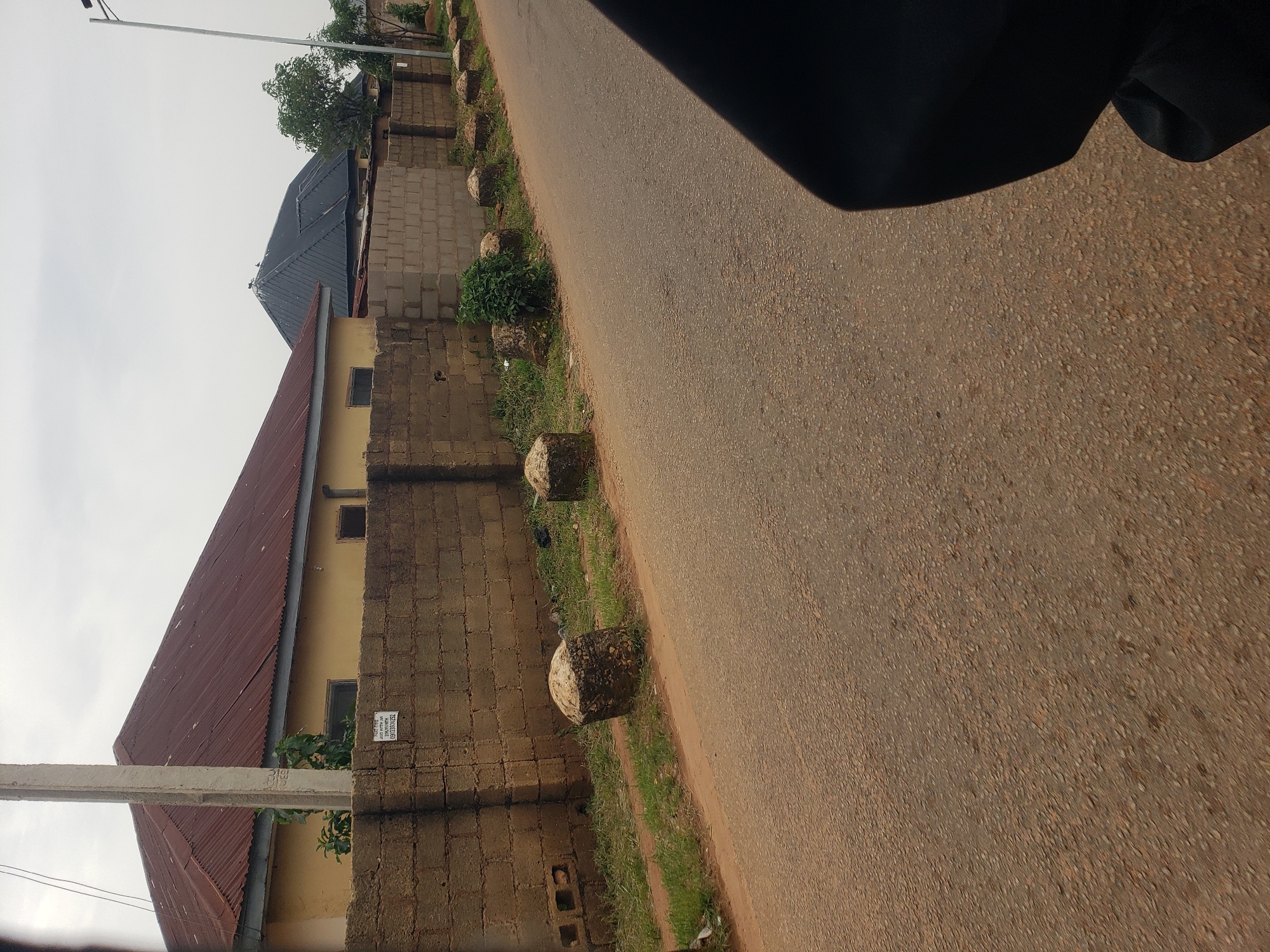 The image depicts a street scene in Bukuru, Nigeria. On the left side of the frame, a pale yellow building with a corrugated red metal roof is visible, partially obscured by a brick wall. The wall is constructed from tan-colored concrete blocks. A sign with text and numbers, possibly a phone number or address, is affixed to the wall. Adjacent to the building, a concrete utility pole stands. In front of the wall, a line of rounded, rough-textured concrete bollards is spaced along the edge of the asphalt road. The road itself appears worn, with some cracks and scattered gravel. Green grass and sparse vegetation grow between the bollards and the wall, and along the side of the road. Further down the street, another building with a dark, angular roof is visible, and a tall, slender metal pole, likely a lamppost, is present on the right. The sky is overcast. A dark, curved object, possibly clothing or a car part, occupies the lower right portion of the image, suggesting the photo was taken from a vehicle or by someone wearing dark attire.