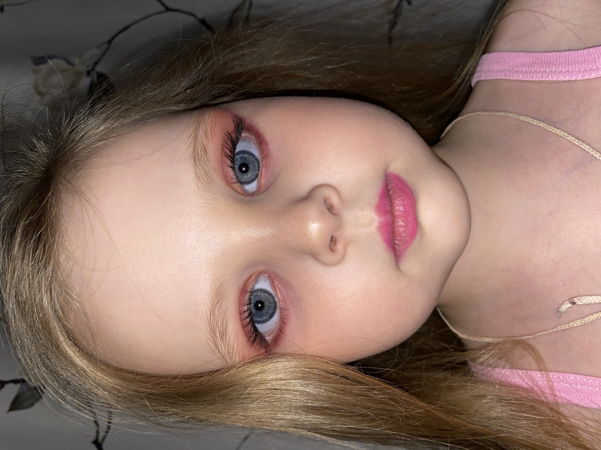 A close-up of a young girl is presented. She has blonde hair, fair skin, and blue eyes. She is wearing pink makeup around her eyes and on her lips. She appears to be lying down on a surface with a patterned background. Her clothing includes a pink top with thin straps and a delicate gold necklace. There is no indication of the location of Votkinsk, Russia, or any city.