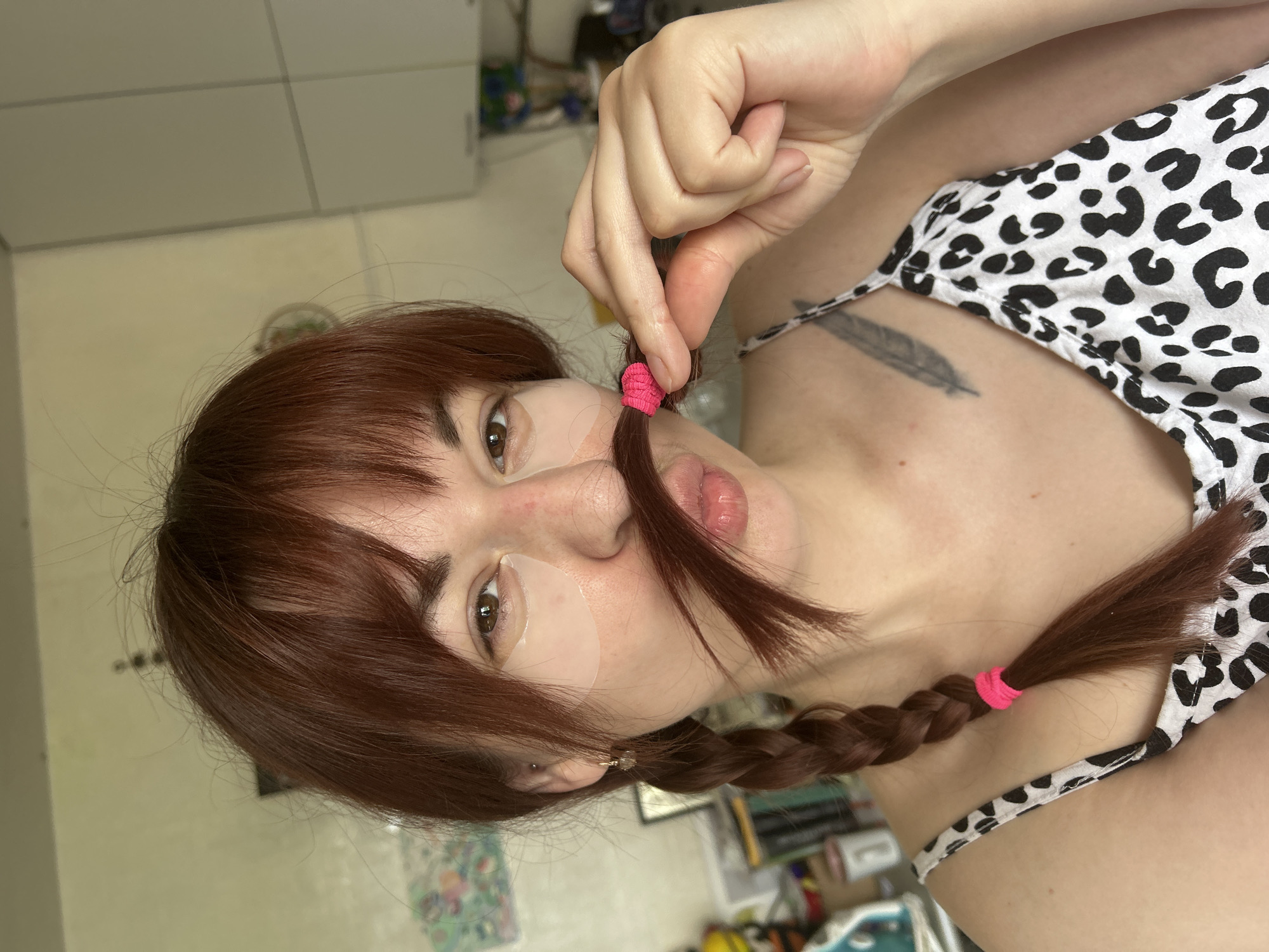A woman is lying down, partially facing the camera. She has auburn hair styled into two braids tied with pink hair ties, and she is wearing a black and white patterned tank top. She has a small bandage or cosmetic patch under her left eye. A feather tattoo is visible on her upper chest. Her hand is holding one of the hair ties from her braid near her face. The background appears to be a room interior with cabinets and various objects, possibly a living space. There is no visible evidence to connect the location to the city of Фоки, Russia.