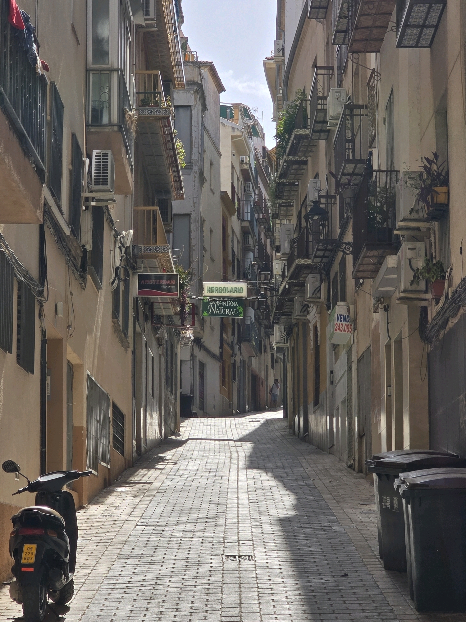 The image captures a narrow street in the city of Jaén, Spain, likely in the historic old town, characterized by its tightly packed buildings and a cobblestone pavement. The perspective looks down the street, which slopes slightly upwards and curves to the right. Sunlight casts sharp shadows across the pavement, indicating it is daytime.
On the left side of the street, a dark-colored scooter is parked facing away from the camera, displaying a yellow license plate with the inscription "C-9 279 BDS". Above the scooter, the building facade features windows with dark-colored shutters. A sign for "AMSTERDAM SEX CENTER" is visible, with red and black lettering. Further up the street, a sign for "HERBOLARIO LA VENTANA NATURAL" is present. Buildings on both sides of the street have multiple stories, with balconies adorned with plants and air conditioning units. Wires and cables are strung between buildings.
On the right side of the street, several dark trash bins are lined up. Further down the street, a person is walking away from the camera. Signs indicating "VEND 243.095" are also visible on the buildings, suggesting properties for sale. The overall impression is of a lived-in urban environment with a mix of residential and commercial establishments.