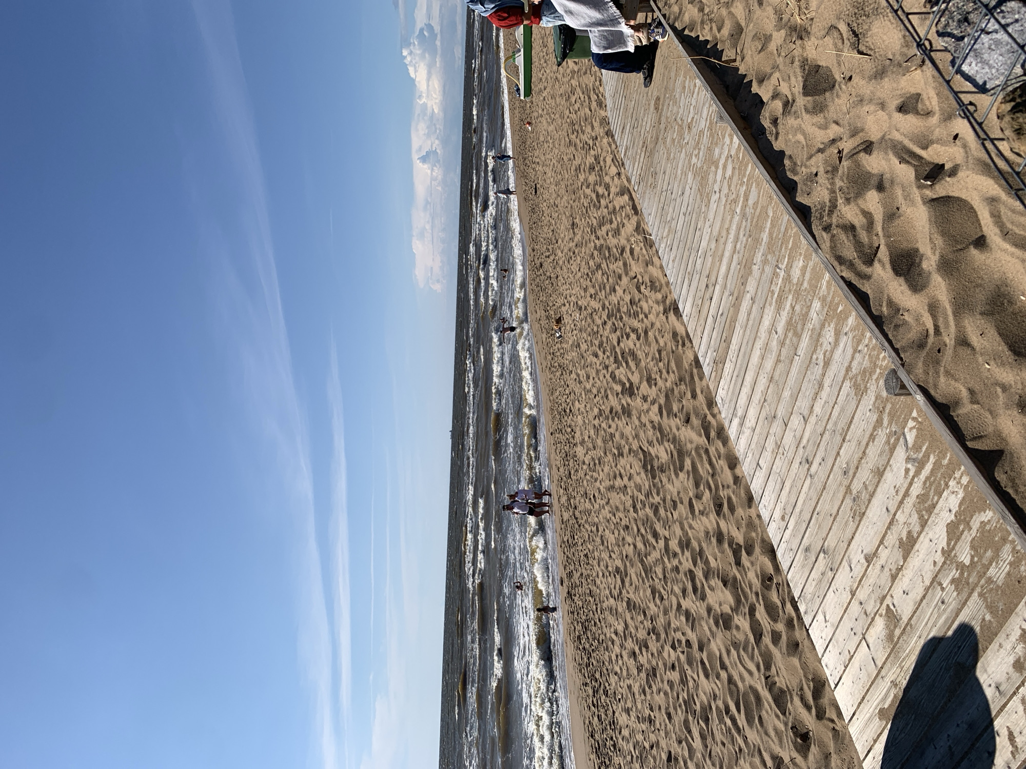 This is a daytime, eye-level, wide shot of a sandy beach in Saulkrasti, Latvia. The scene is dominated by a bright blue sky with scattered white clouds and a choppy, brown body of water that appears to be the Baltic Sea. A wooden boardwalk runs diagonally across the foreground, leading towards the beach and the water. Several people are visible on the beach and in the water, engaged in various activities such as walking, playing, and swimming. A green pedal boat is situated on the sand near the boardwalk, and a person is seated on a bench to the right of the frame. The overall impression is one of a lively summer day at the seaside.