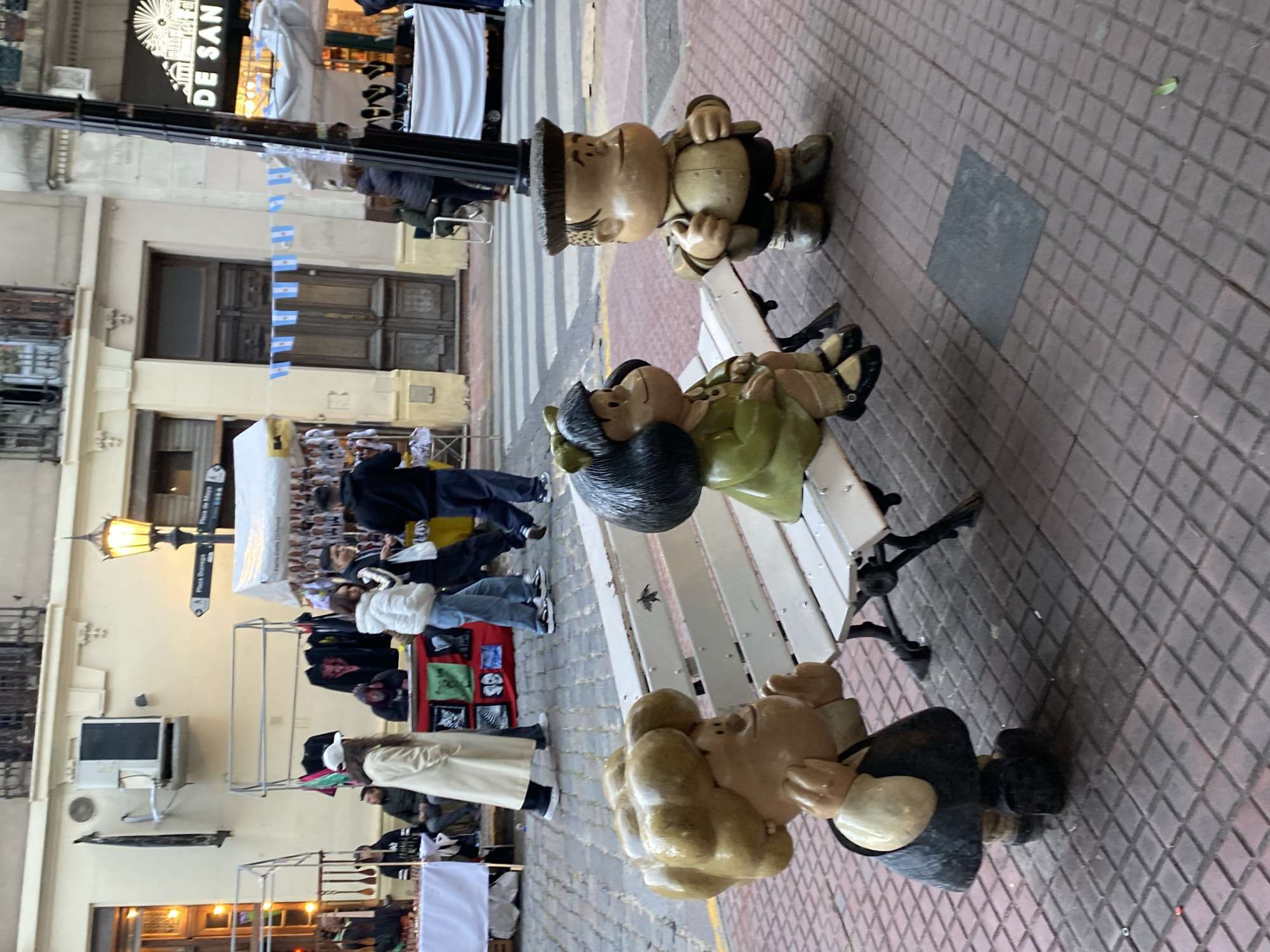 The image shows a public art installation depicting the Argentine comic strip characters Mafalda, Manolito, and Felipe sitting on a white bench on Calle Defensa in Buenos Aires, Argentina. Calle Defensa is paved with red bricks. A street fair is set up along the street with vendors selling various items, and people are walking around browsing the stalls. The street is lined with buildings, some with awnings. There is a street sign indicating Plaza de Mayo and Defensa. The sky is overcast.