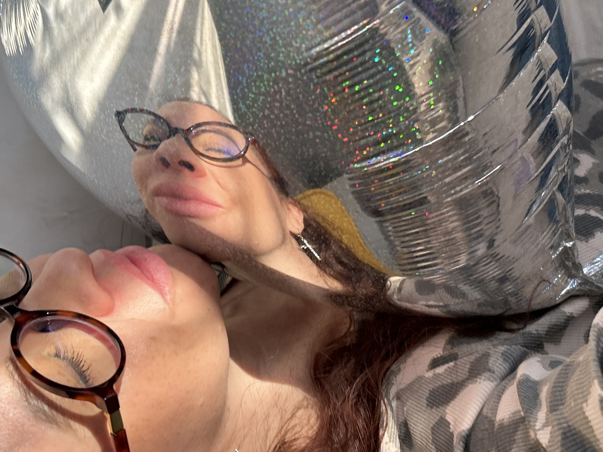 This close-up image features two individuals, likely women, bathed in strong, direct sunlight. In the foreground, on the lower left, is the profile of one person's face, eyes closed, with pronounced eyelashes visible through brown tortoise-shell glasses. Their lips are full and pink. Partially obscured behind and to the right, the second person's face is seen looking upwards, wearing similar glasses, with a slight smile on their red lips. This individual has reddish-brown hair that cascades around their head and neck.
Dominating the background and upper right is a large, highly reflective silver object, most likely a balloon. Its surface is adorned with holographic glitter, scattering iridescent rainbow speckles in the bright sunlight. Another softer, reflective silver surface is visible on the far left. The harsh light creates sharp shadows and brilliant highlights, suggesting a sunny day, possibly indoors near a window. A patterned fabric, possibly grey and white camouflage or plaid, can be seen on the right side of the frame, hinting at clothing or bedding. No text is visible. The scene conveys a relaxed and possibly celebratory mood due to the festive balloon.