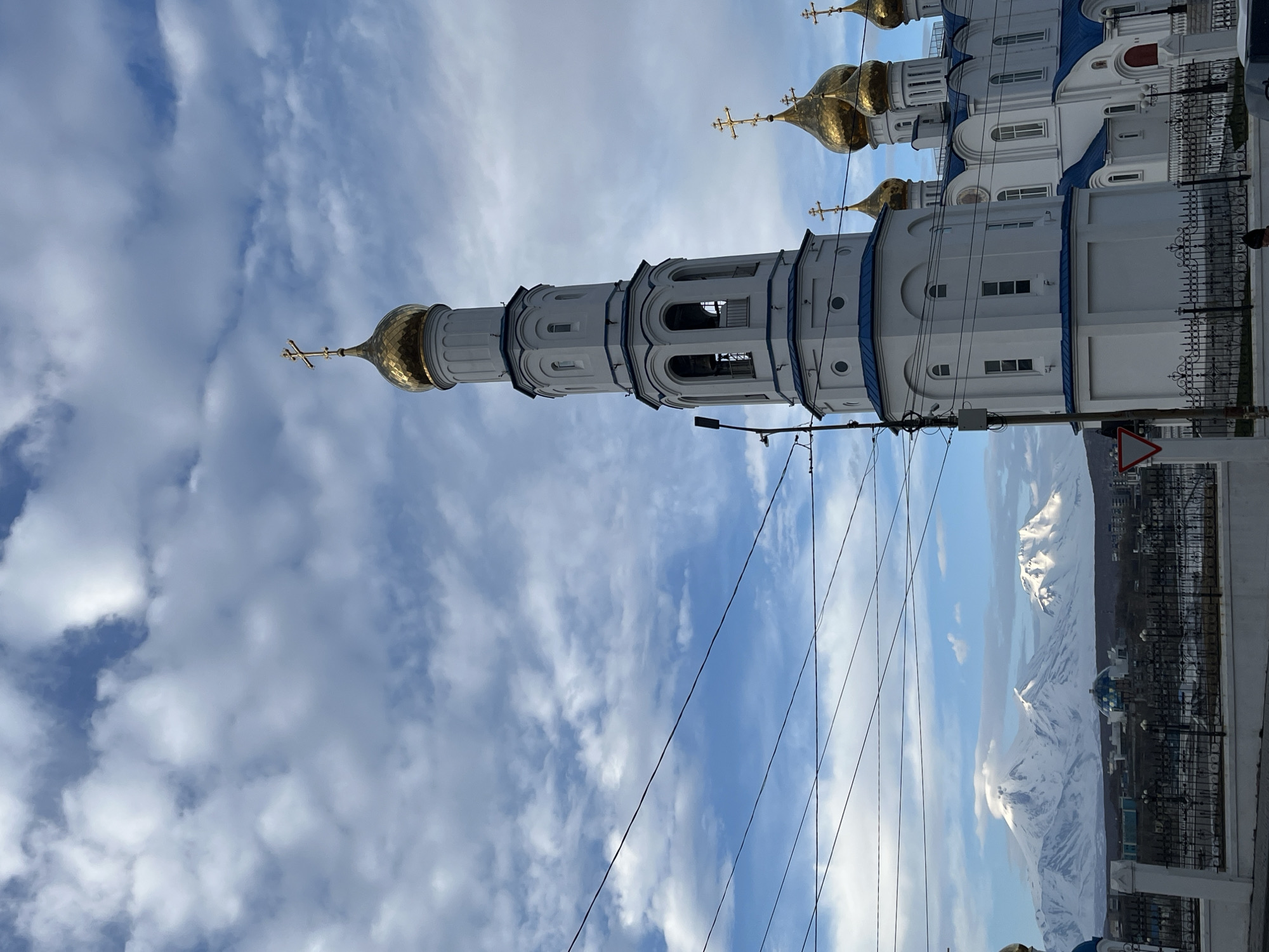 The image depicts the Holy Trinity Cathedral in Petropavlovsk-Kamchatsky, Russia, against a backdrop of cloudy sky and distant snow-capped mountains. The cathedral's white facade with blue trim and golden domes is prominently featured. Power lines are visible in the foreground, cutting across the view of the sky and mountains. A street sign is present on the right side of the image. The photograph is taken from a low angle, emphasizing the height of the cathedral. The presence of a metal fence suggests the location is a public area.