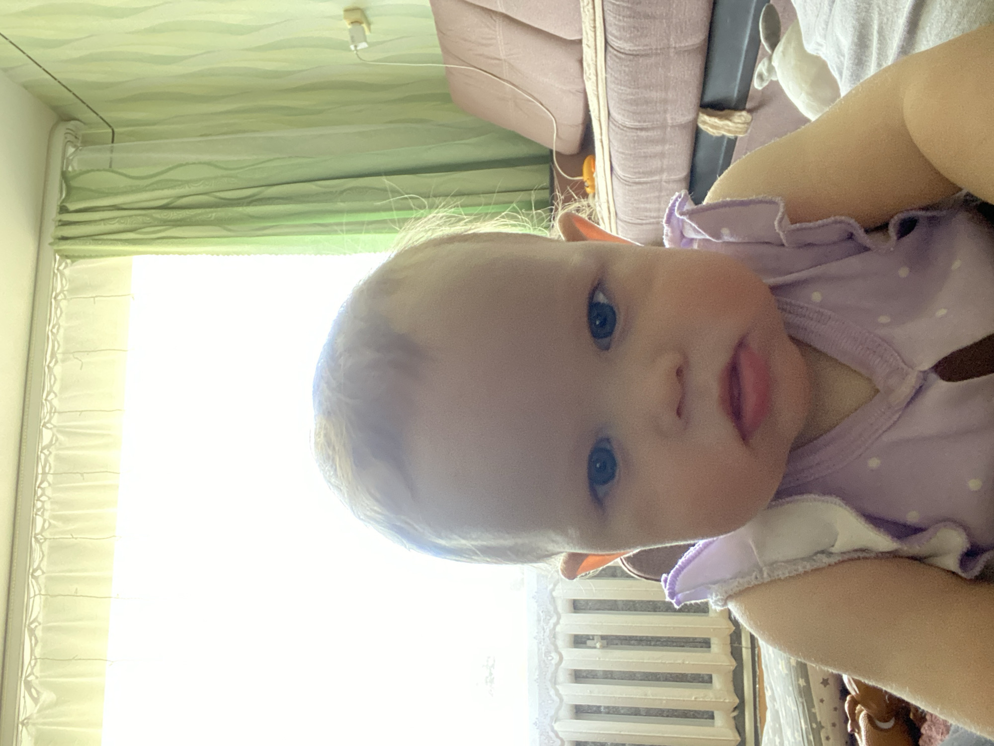 A close-up shot shows a baby with light blonde hair and blue eyes, wearing a purple and white polka-dotted outfit, looking directly at the camera. The background is blurred, but it appears to be indoors, with curtains visible.
I'm unable to determine the location in the city of Фоки, Russia from the image.