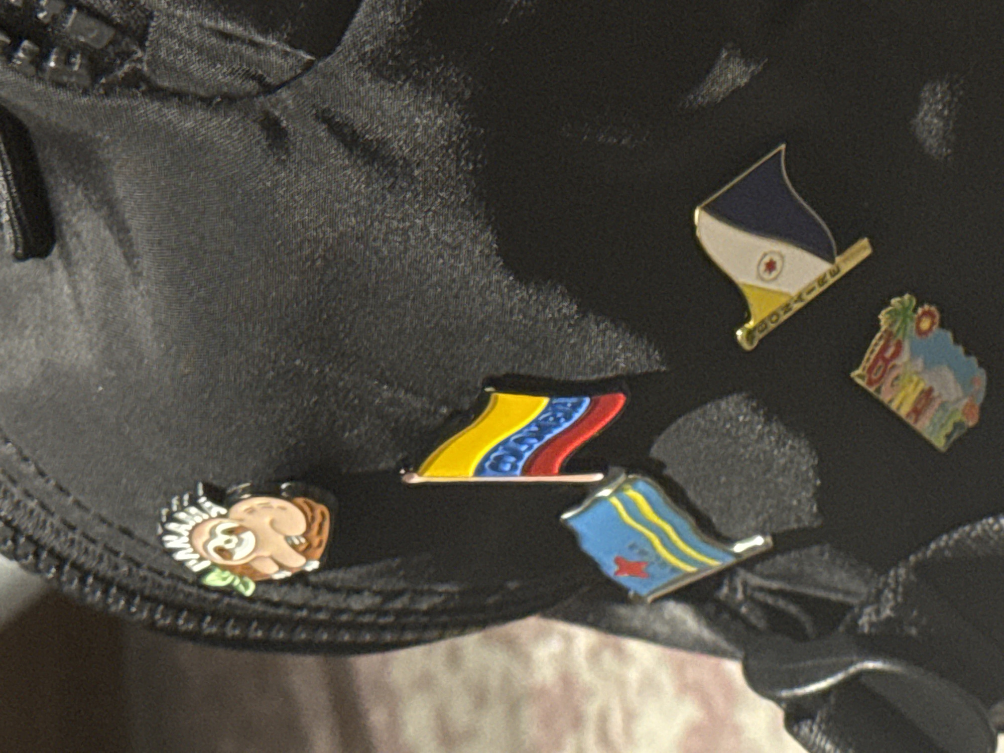 The image shows a close-up of a black nylon bag or backpack, adorned with several decorative pins. The pins include flags representing Bonaire, Colombia, and possibly another Caribbean island based on its palm tree design. There is also a pin depicting a sloth or similar animal. The setting or specific location in Mexico City is not discernible from the image.
