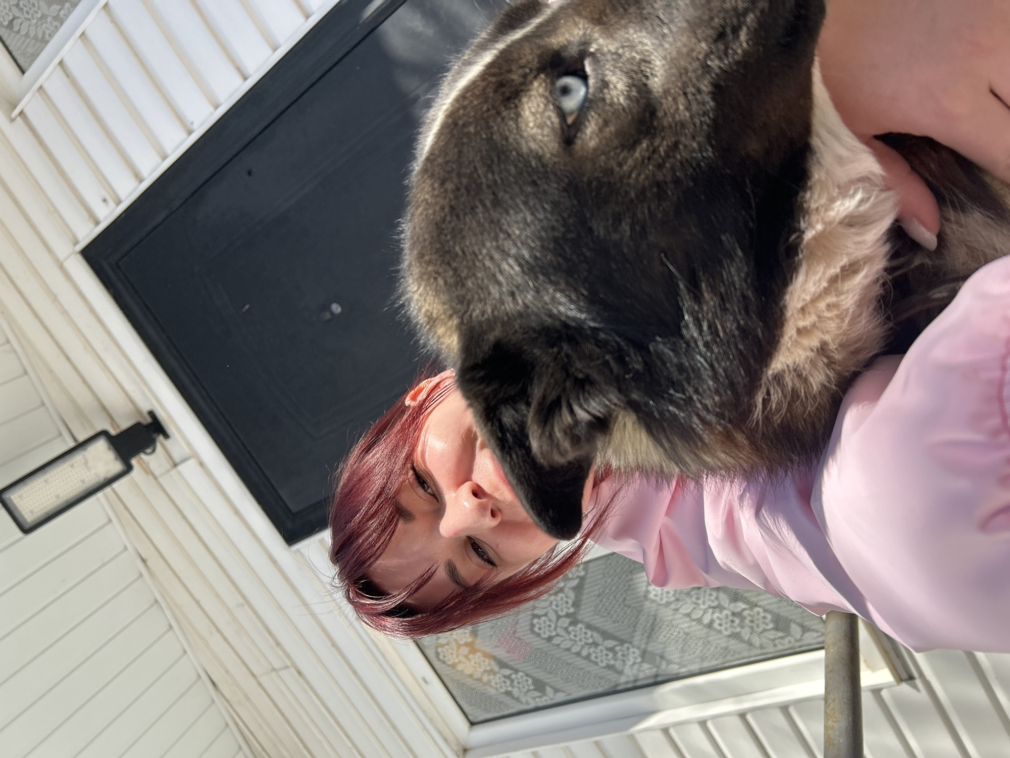 A low-angle, portrait-style photograph shows a woman holding a dog in her arms outside. The woman has red hair, and she is wearing a pink jacket. The dog, which is the more prominent subject in the shot, has dark fur with white patches around its neck and blue eyes. They are both looking downward, seemingly toward the camera.
Behind them, a white, wood-paneled building structure with a black door and a rectangular outdoor light fixture is visible. The photograph appears to have been taken in natural light.
It is not possible to determine the exact location within the city of Фоки, Russia from the provided image.