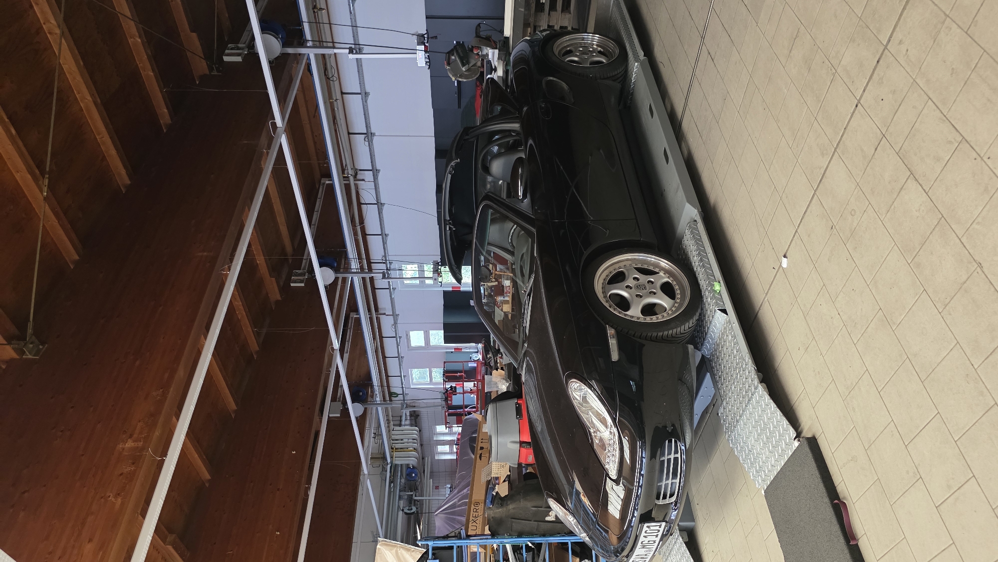 The image depicts a black convertible sports car, identified by its license plate as "SW A-MG 101", positioned on a car lift within a workshop. The car has its roof partially open. The workshop environment features a wooden ceiling with exposed beams and industrial lighting fixtures. Various tools and equipment, including a red tool cart and what appears to be a vacuum cleaner, are visible in the background, along with shelving units and stacked boxes. The floor is tiled. The overall scene suggests a repair or maintenance setting.