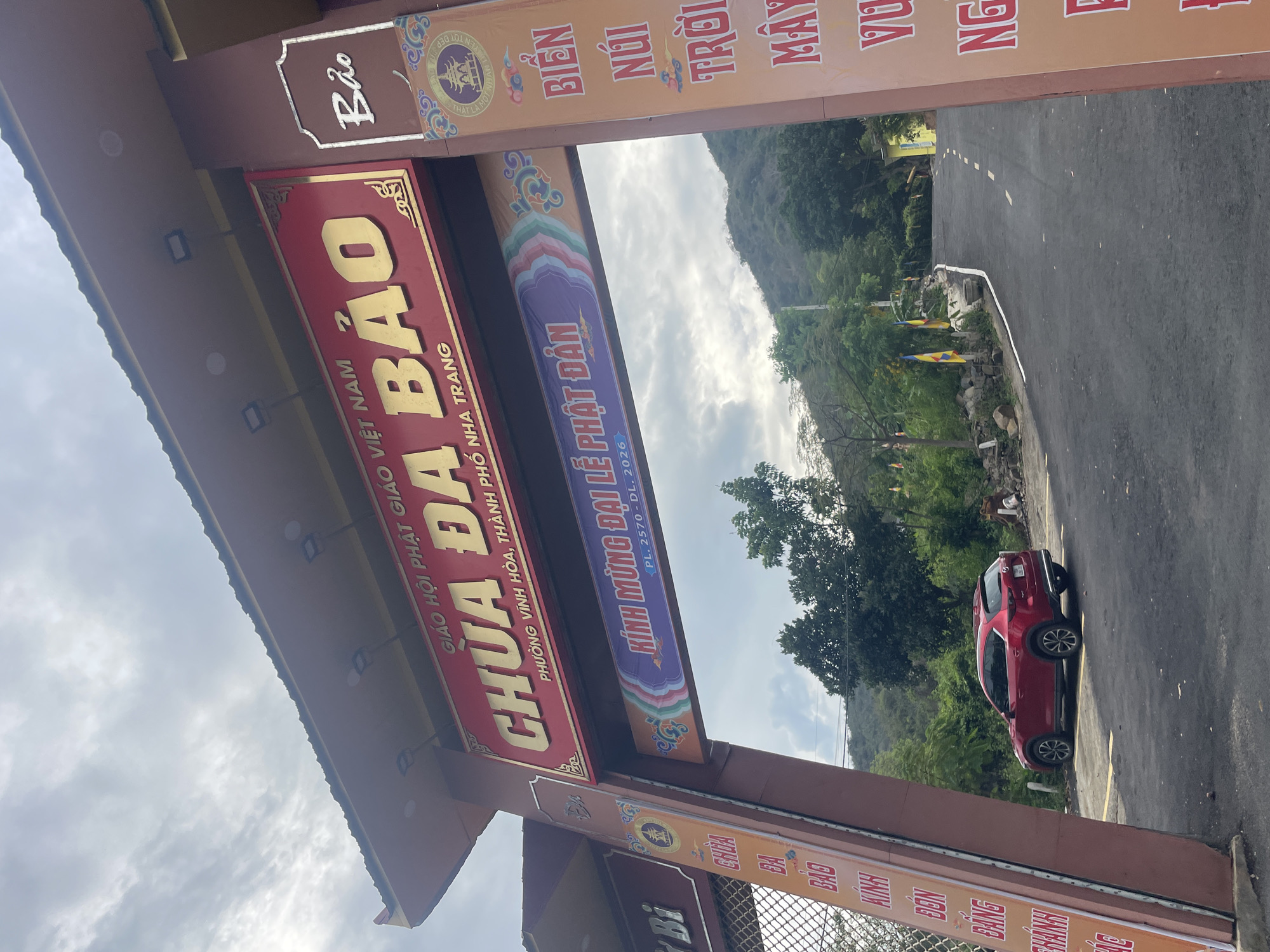 The image displays a grand entrance gate or archway, likely belonging to a temple, under a cloudy daytime sky. The perspective is looking upwards and through the archway.
The prominent archway features multiple signs and banners in Vietnamese. A large red sign with gold lettering at the top of the arch reads: "GIÁO HỘI PHẬT GIÁO VIỆT NAM" (Buddhist Sangha of Vietnam) and "CHÙA ĐA BẢO" (Da Bao Pagoda), followed by the address "PHƯỜNG VĨNH HÒA, THÀNH PHỐ NHA TRANG" (Vinh Hoa Ward, Nha Trang City). Below this, a colorful banner declares: "KÍNH MỪNG ĐẠI LỄ PHẬT ĐẢN" (Celebrating the Great Buddha's Birthday Festival) with the dates "PL. 2570 - DL. 2026" (Buddhist Era 2570 - Common Era 2026), indicating a future celebration. Other fragmented text visible on surrounding banners includes "Bảo", "BIỂN", "NÚI", "TRỜI", "MÂY", "VU", "NG", "BÁNH", "Bi", "CHDA", and "BA", possibly referencing local elements or further details about the temple or region.
Beyond the archway, an asphalt road is visible, curving gently to the right. A red SUV is parked on the right side of this road. The area surrounding the road is verdant with numerous trees and foliage, leading to mountains in the distance. The setting is in a natural, somewhat hilly or mountainous environment, characteristic of Bac Nha Trang Ward, Vietnam. The weather appears overcast, and the time of day is daytime, with no individuals visible in the scene. The overall impression is of an entrance to a significant religious site, preparing for or celebrating a major Buddhist festival.