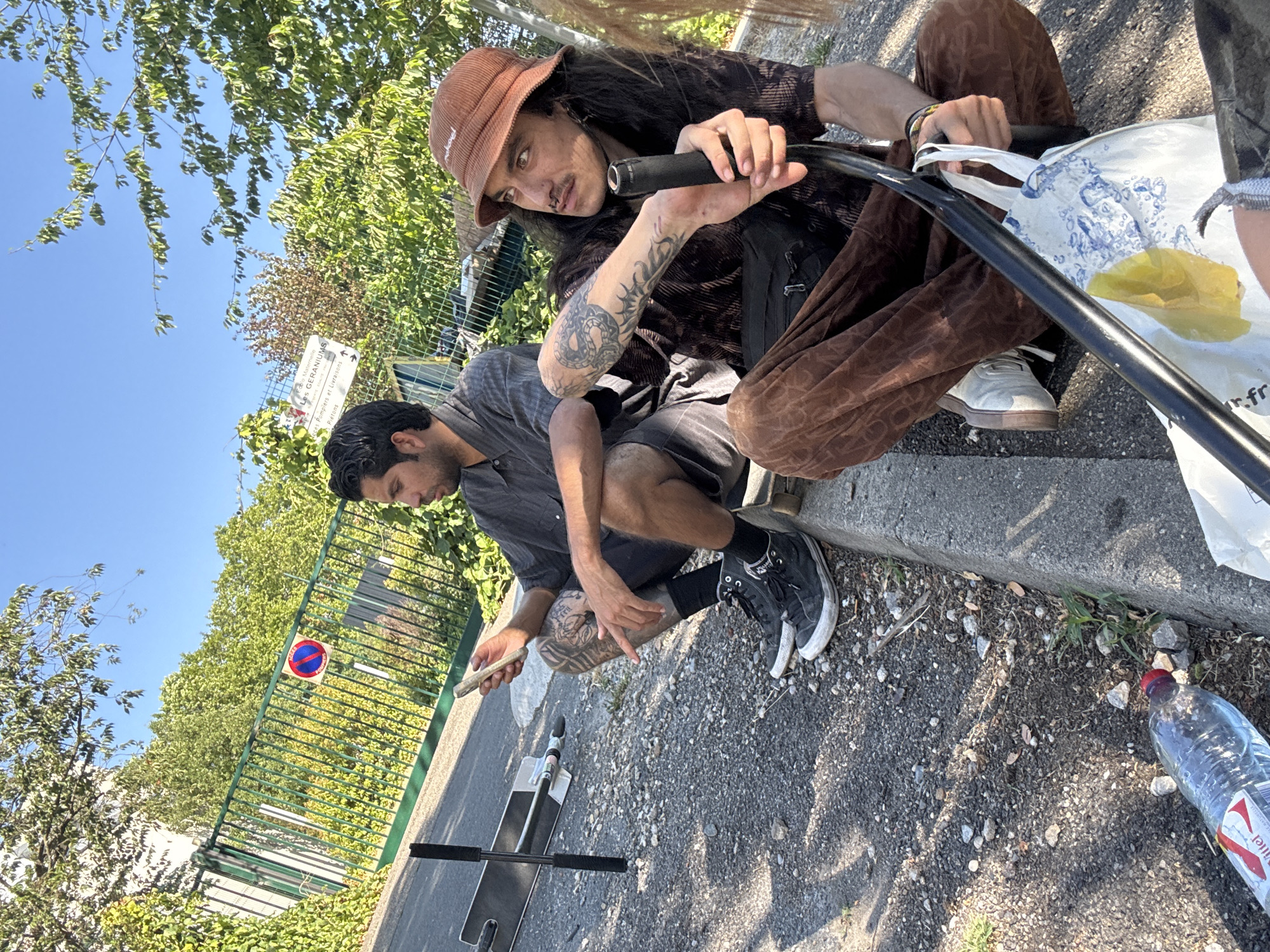 Two young men are sitting on the side of a road near a metal fence in Lyon, France. The man on the left, wearing a grey shirt and black shorts, is holding a smartphone in his hand. He has tattoos on his left arm and leg. He is wearing black high-top sneakers. Next to him is a scooter. The man on the right is wearing a brown bucket hat and a patterned brown shirt. He has long dark hair and tattoos on his arm. He is holding a black object, possibly part of a bicycle or scooter, and a white bag with blue and white patterns. He is wearing brown pants and white sneakers. A plastic water bottle is on the ground near the curb. The scene is outdoors under a clear blue sky with trees and greenery in the background. There are traffic signs visible on the fence.