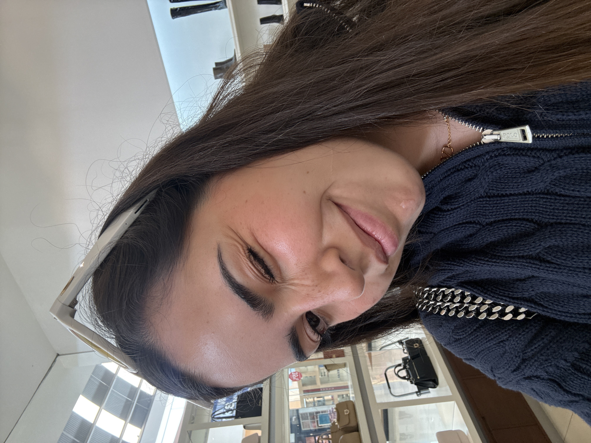 A woman is posing indoors. She has long brown hair, dark eyebrows, and is wearing sunglasses pushed up on her forehead. She is wearing a dark blue cable-knit sweater with a zipper and a silver chain. The zipper pull says "POLO." A gold necklace with a pendant is visible.
Through the window behind her, storefronts are visible. The location appears to be San Francisco Ocotlán (Ocotlán), Mexico. Manicure chairs, displays, and merchandise are inside the building.