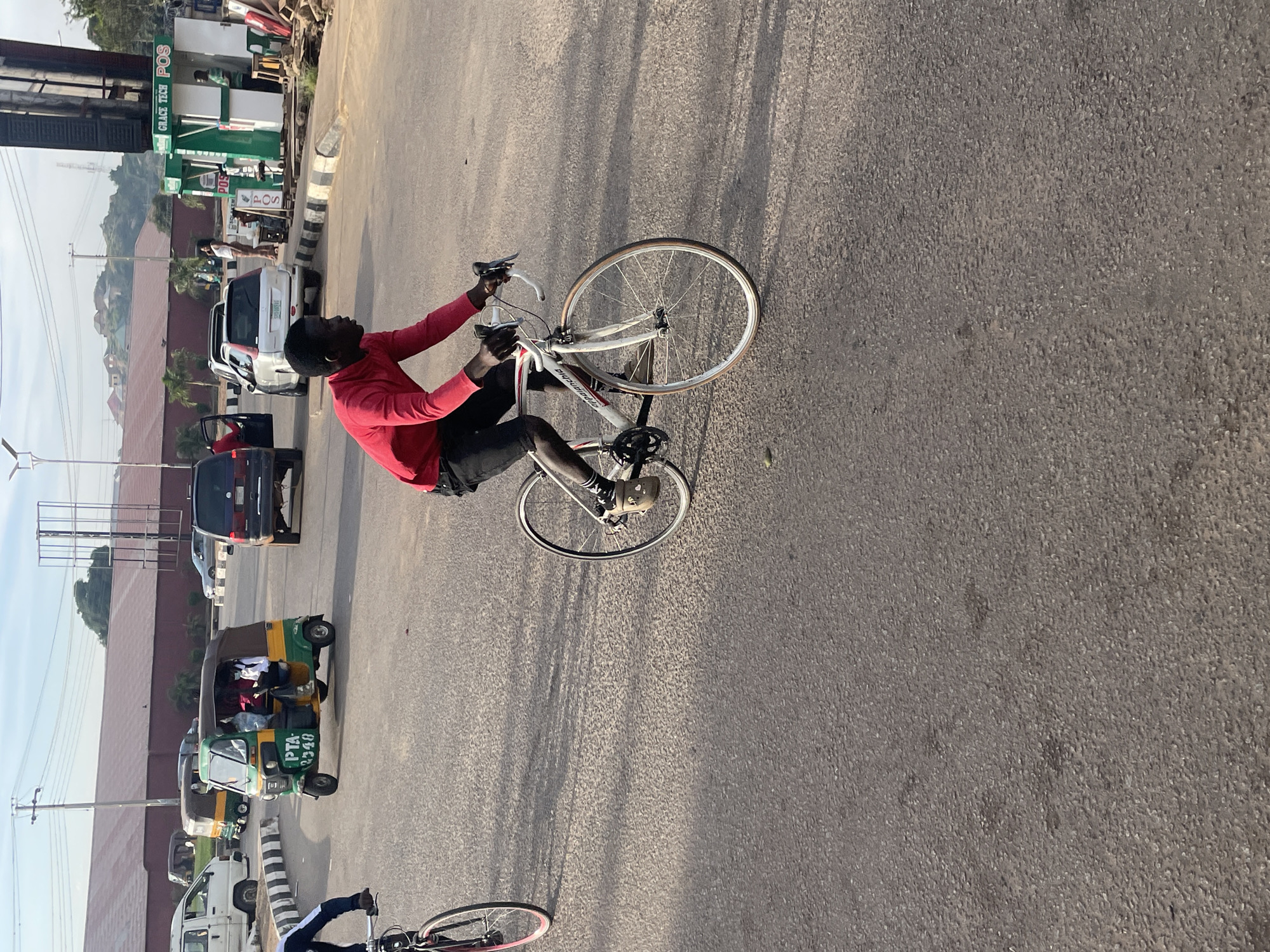 The image captures a bustling street scene in Jos, Nigeria, likely during daylight hours given the strong shadows. The foreground is dominated by a person riding a white bicycle with red handlebars. This individual is wearing a red long-sleeved shirt and dark shorts, with a visible black sock and a light-colored shoe on their right foot. Their left leg is not fully visible. The bicycle has thin wheels and a visible chain and gears. To the left, partially visible, is the front wheel and handlebars of another bicycle, suggesting multiple cyclists are present.
In the mid-ground, a green and yellow auto-rickshaw, commonly known as a Keke Napep, is moving. It displays the license plate "PTA 2540" and appears to be carrying passengers. Behind the Keke Napep, a dark-colored SUV is also on the road. Further back, a white SUV is parked with its driver's side door open. To the right of the white SUV, a roadside kiosk with green and white signage reads "GRACE TECH POS," indicating a financial service point. A person in a light-colored shirt stands near the kiosk, and another individual in a green shirt is visible inside.
The background features a large, red-brown building with a corrugated metal roof, possibly a warehouse or factory. Beyond that, a hilly landscape with scattered buildings and vegetation is visible under a clear, pale blue sky. Power lines and streetlights punctuate the urban environment. The road surface is asphalt, showing signs of wear and tear, with visible tire marks and scattered debris. The overall impression is one of everyday urban activity and transportation in Jos.