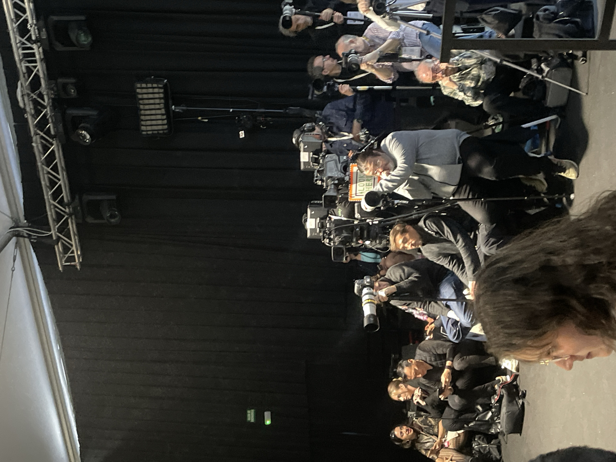 The image depicts a group of people, possibly photographers or camera operators, positioned in rows with their equipment. The scene appears to be indoors, likely in a media or press area. The background is dark, with black drapes or curtains. Equipment, including cameras with large lenses, tripods, and lighting rigs, is prominent.
Without further information, pinpointing the exact location in Barcelona is impossible. The image provides no clues to identify the specific venue.
