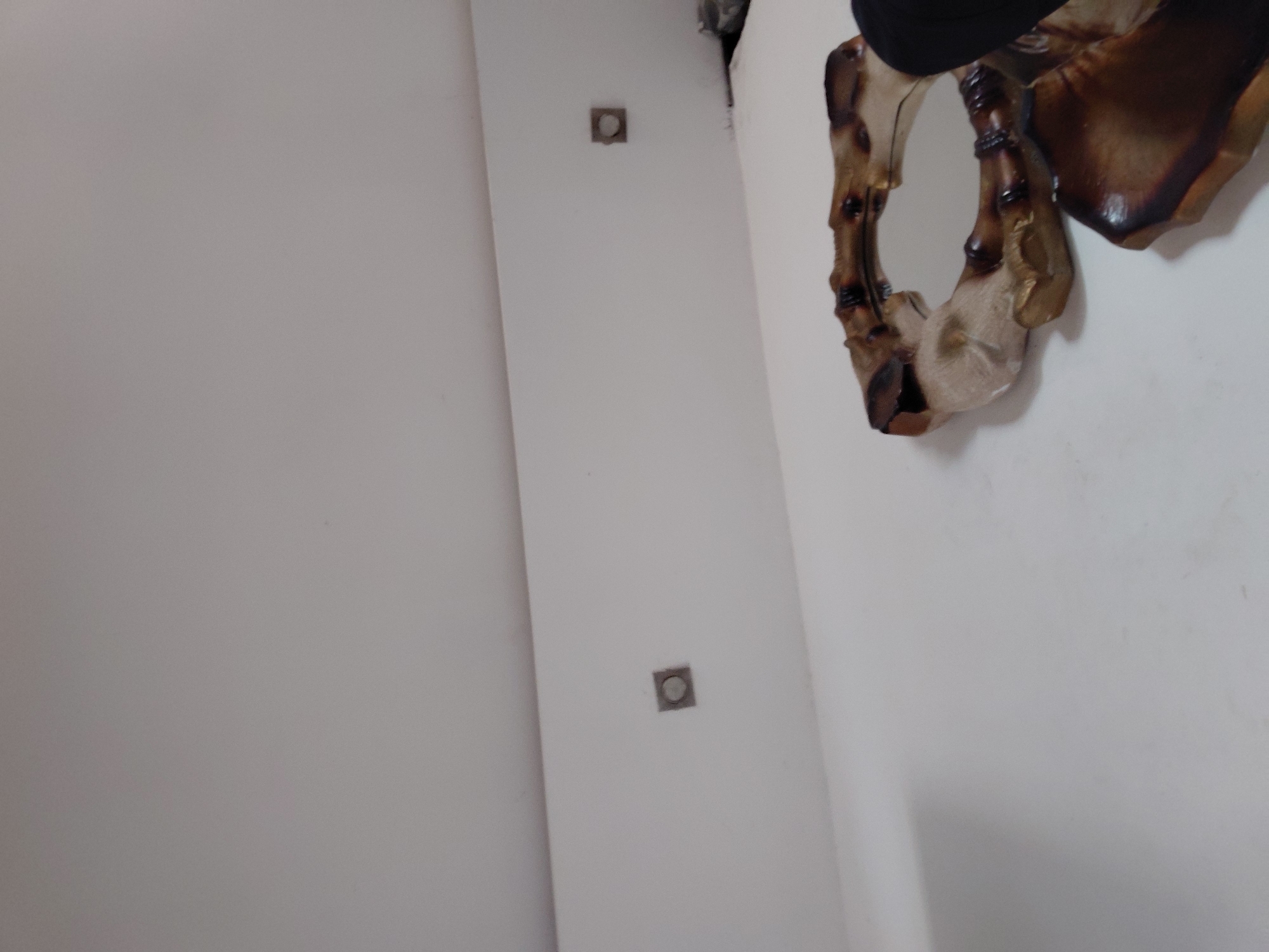 This media file, captured in Santa Ana, Venezuela, presents an oblique view of an indoor setting, focusing on architectural elements and a decorative object. The scene is dominated by plain white walls or ceiling surfaces, suggesting a minimalist or modern interior.
On the right side of the frame, a distinct decorative mirror or art piece is partially visible. Its frame is irregularly shaped, featuring a dark brown, almost burnt or heavily stained material, contrasting with lighter, bone-like or beige sections. The design is organic, appearing sculptural with flowing lines and a texture reminiscent of polished wood or resin. A dark, undefined object is partially visible above the mirror, possibly a ceiling fixture or part of the room's upper structure.
On the left side, two identical square-shaped fixtures are recessed into the white surface, positioned vertically one above the other. Each fixture contains a small circular element in its center, suggesting they are either recessed lighting fixtures, smoke detectors, or sensors. The image is taken from a low angle, looking upwards towards a corner or a prominent vertical feature of the room. No activity is taking place, and no people or visible text are present. The lighting appears even, indicating an indoor environment without strong directional light sources.