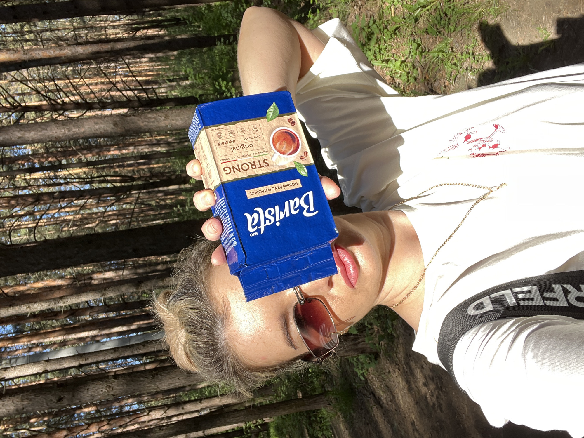 A person lies on their back outside in a forest-like area, holding a blue box of Barista brand coffee over their forehead and sunglasses. The box is labeled "STRONG" and "original" in English, with Russian text below. The person wears a white shirt with a cartoonish design on the chest and a black strap visible at the shoulder with the name "RFELD" on it. Trees are visible in the background. The location is Chaikovsky, Russia.