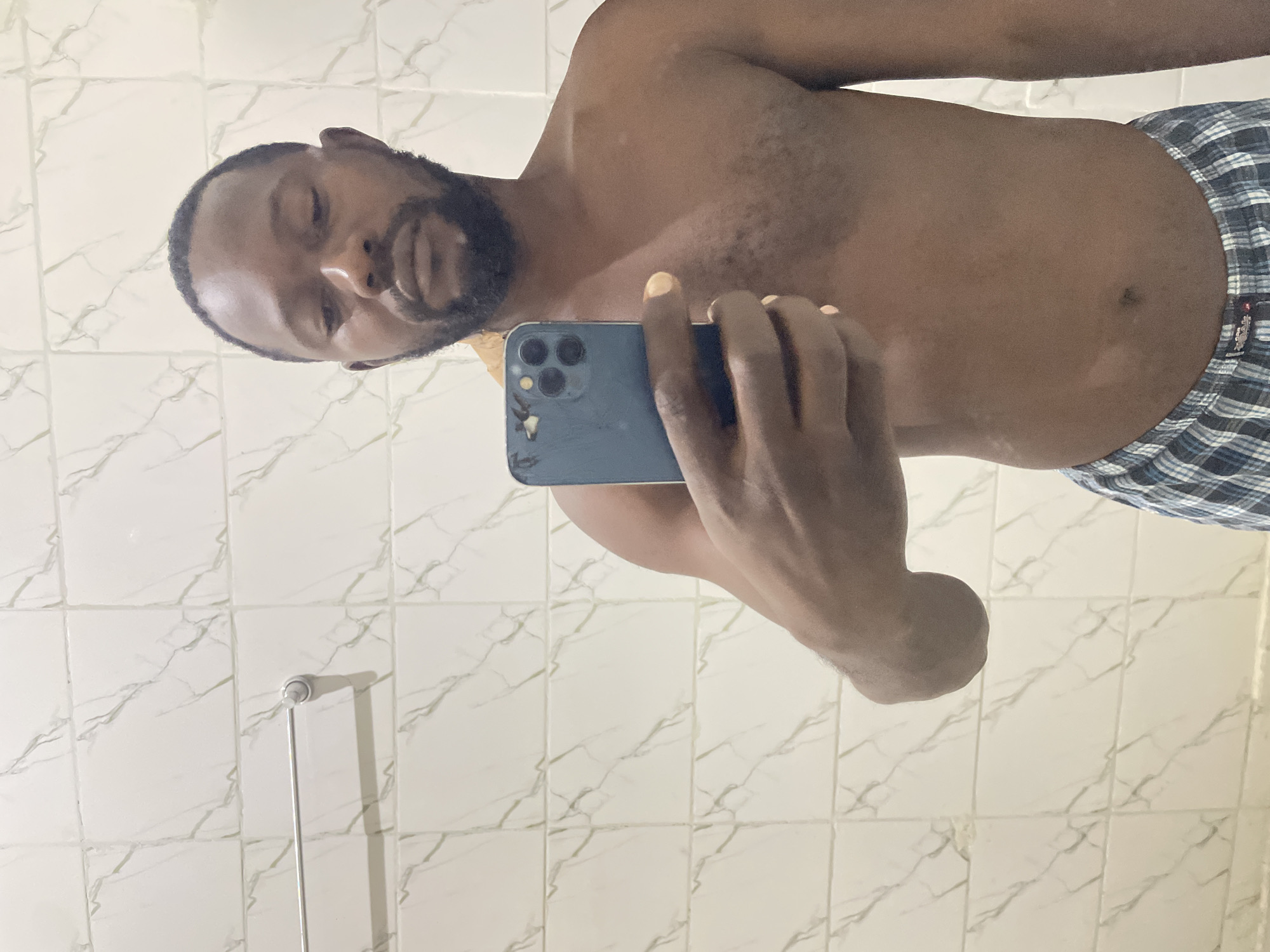 A shirtless Black man with short dark hair and a beard takes a mirror selfie in a tiled bathroom. He is holding a blue smartphone in his right hand. The bathroom walls are covered in white square tiles with a marble pattern. A silver towel rack is mounted on the wall behind him. He is wearing blue and white plaid boxer shorts.