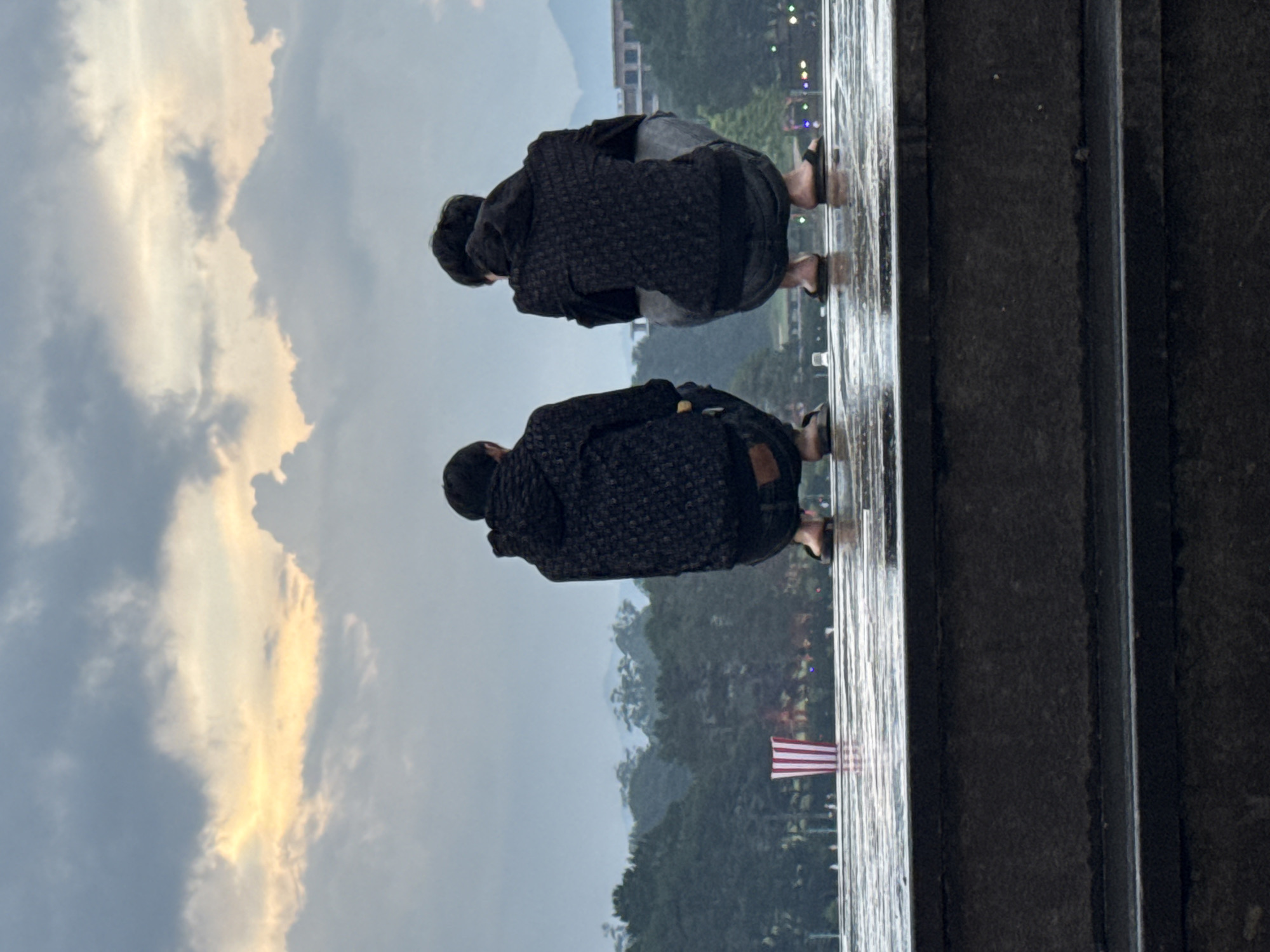 Two people are crouching on a wet, reflective surface, possibly a bridge or platform. They are both wearing dark clothing and sandals. The background shows a landscape with trees, a body of water, and a flag, possibly the American flag. The sky is cloudy with some patches of light. The location is likely Da Lat, Vietnam.