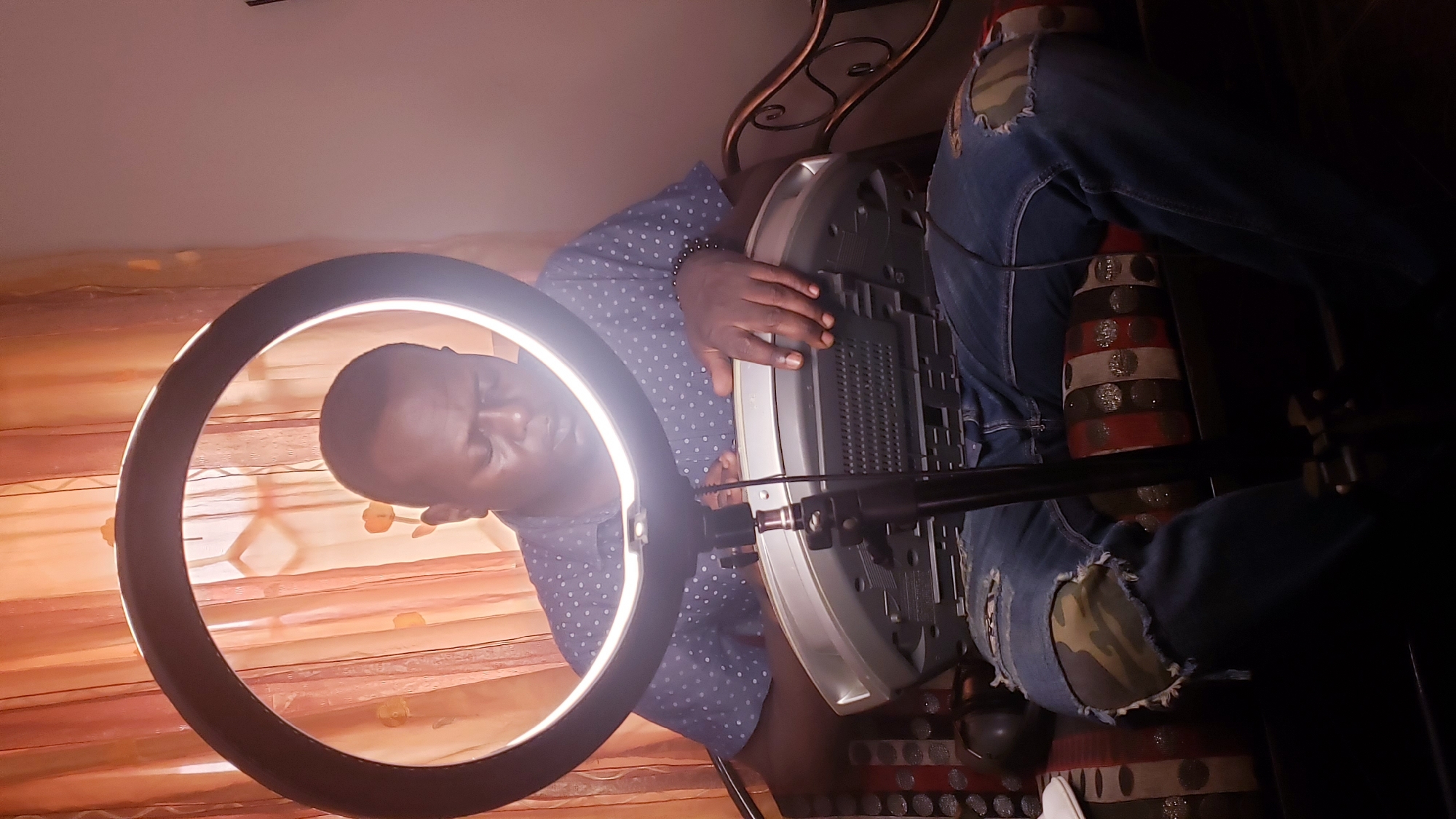 A dark-skinned man sits on a couch in front of a sheer, peach-colored curtain. He is wearing a blue shirt with white polka dots and ripped blue jeans with camouflage patches on the knees. A large ring light is positioned in front of him, illuminating his face and the silver boombox he is holding in his lap. The boombox is partially obscured by his arms and the ring light stand. The couch has a striped pattern with colors of red, white, and black, and appears to be made of metal with decorative scrollwork on the right side. The overall lighting is dim, with the ring light creating a bright focal point on the man and the boombox. The setting appears to be indoors, possibly a living room, in Narayi, Nigeria.