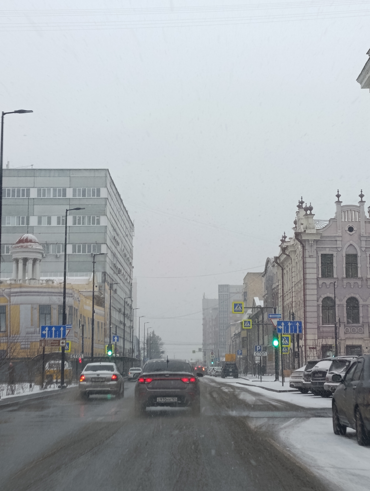 The image depicts a snow-covered street in Krasnoyarsk, Russia, under a dull, overcast sky with light snowfall. It appears to be daytime, with a muted color palette dominated by greys, whites, and muted building tones. The road is wet and slushy, indicating ongoing or recent precipitation.
In the foreground and mid-ground, several cars are visible. A red car, identified by its license plate "c970hx 124", is centrally positioned, followed by a silver car with a partially visible license plate "в011ум 124". Other vehicles are parked along the right side of the street, many covered in snow. A green traffic light is visible on the right, suggesting cars are either stopped or proceeding.
The street is flanked by buildings of diverse architectural styles. On the left, a tall, multi-story grey building with numerous rectangular windows stands behind a more traditional, yellow building featuring a classical columned portico topped with a reddish dome. On the right side of the street, a row of ornate, older buildings with intricate facades, decorative rooftops, and arched windows line the street. Their colors range from light purple-grey to cream.
Numerous streetlights, traffic signals, and road signs are scattered throughout the scene. Visible signs include yellow pedestrian crossing signs, blue directional signs with white arrows (e.g., "↑↑↑", "←", "↑", "←"), and a white sign with Cyrillic text reading "СТОР" (likely "СТОП" for stop) next to a "P" for parking. The general setting is an urban environment experiencing typical winter weather conditions, with snow on the ground and building ledges. No specific people are prominently visible, and the overall mood is quiet and somewhat subdued due to the weather.