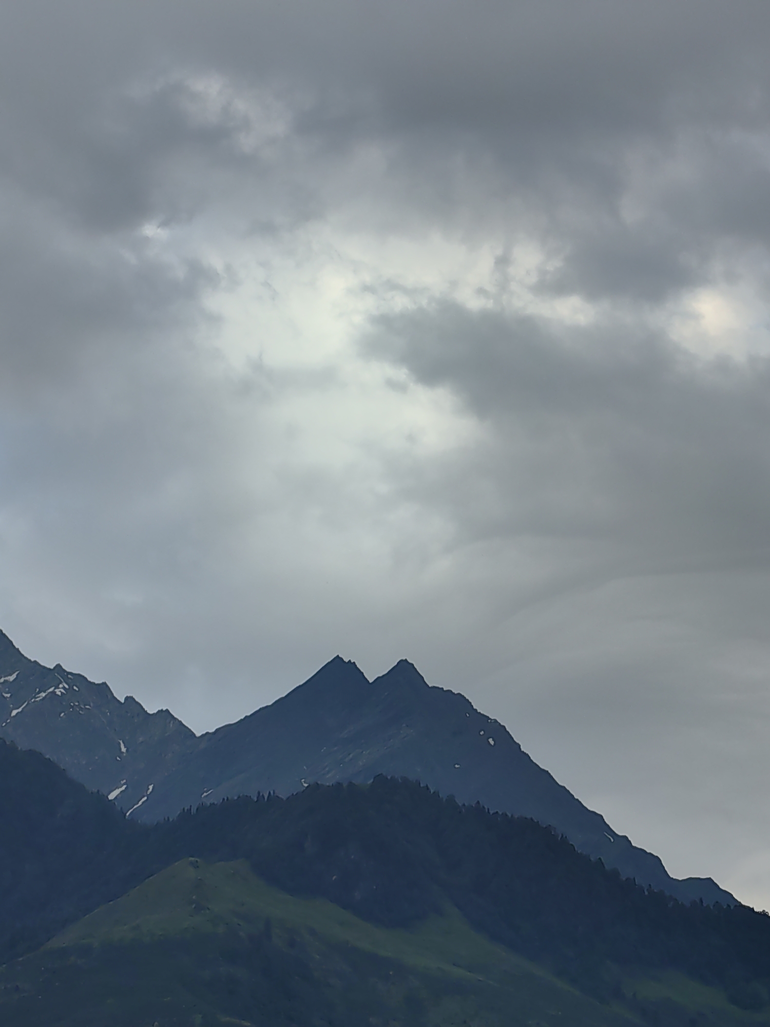 The image depicts a mountainous landscape under a heavily overcast sky. Dark, jagged peaks dominate the foreground, with some patches of snow visible on the upper slopes. A forested slope, rendered in deep shadow, lies in front of the highest peaks. A rounded, green hill occupies the bottom portion of the frame, suggesting a lower elevation within the mountain range. The sky is filled with swirling grey clouds, creating a dramatic and somber atmosphere. This scene is likely located in or near Vashisht, India, a town known for its mountainous terrain. There are no people or animals visible in the image.