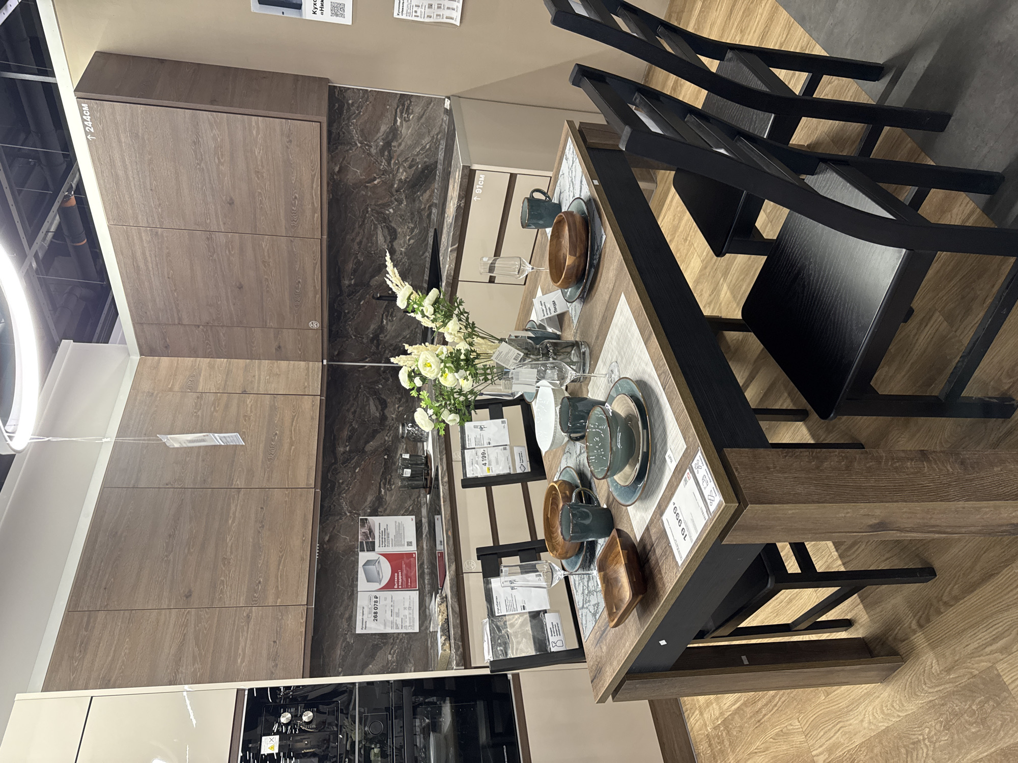 The photo shows a kitchen display in a store. The kitchen cabinets are light brown wood with a marble-patterned backsplash. There is an oven built into the lower cabinets. On the table are dishes, cups, bowls, and a vase with white flowers. There are two black chairs pushed in at the table. There are price tags and information sheets on the table and cabinets, with some Russian text visible. The store is located in Izhevsk, Russia.