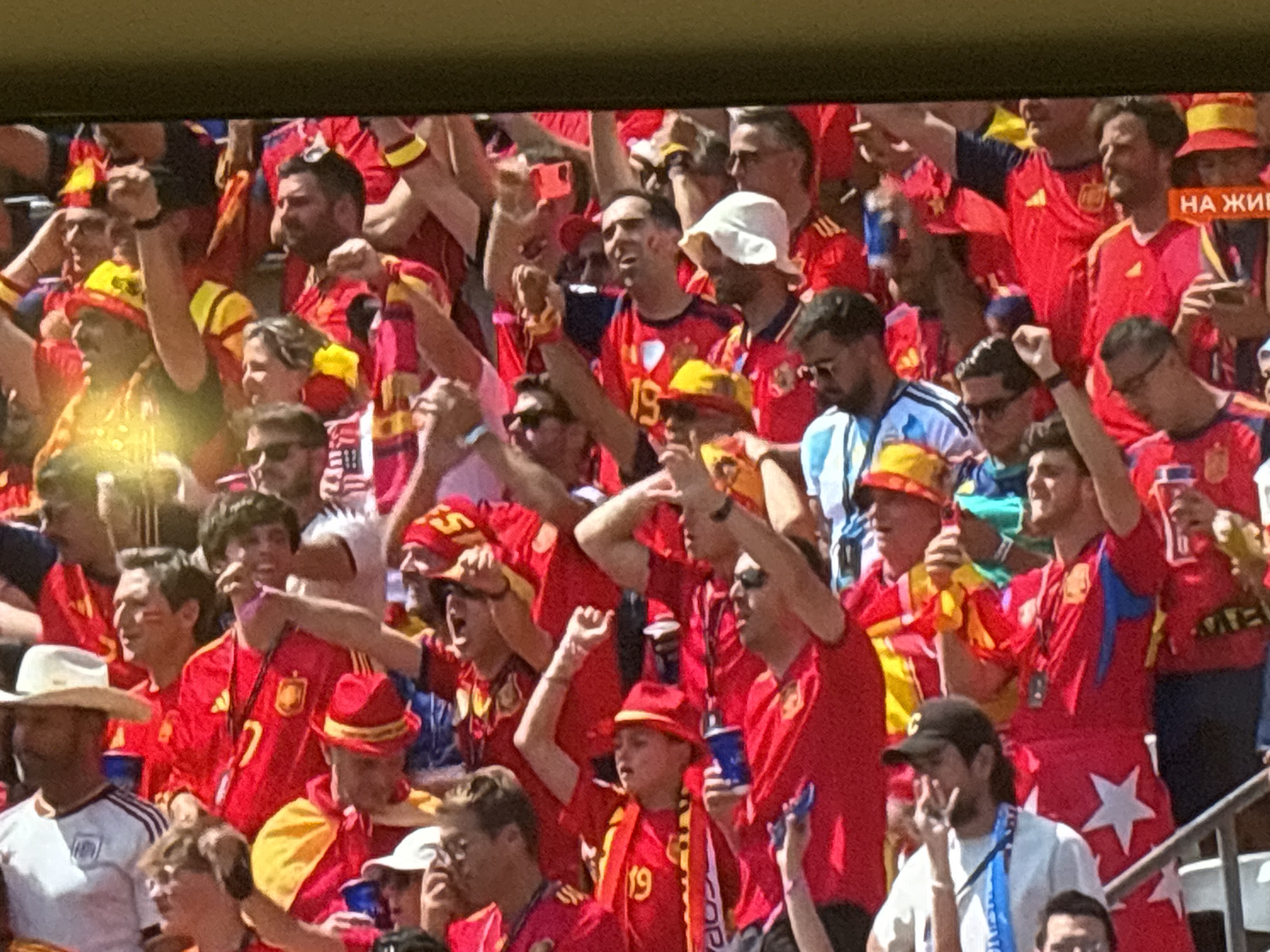 The image shows a crowd of people at a sporting event, likely a soccer match, given the fan attire and energetic atmosphere. Most individuals are wearing red and yellow, suggesting they are supporters of a team associated with those colors, possibly Spain. Many are raising their fists or waving, indicating excitement or celebration. Some are holding up phones, likely recording or taking pictures. The background shows a stadium setting with blurred seating. There is no information in the image to suggest any connection to Burgas, Bulgaria.