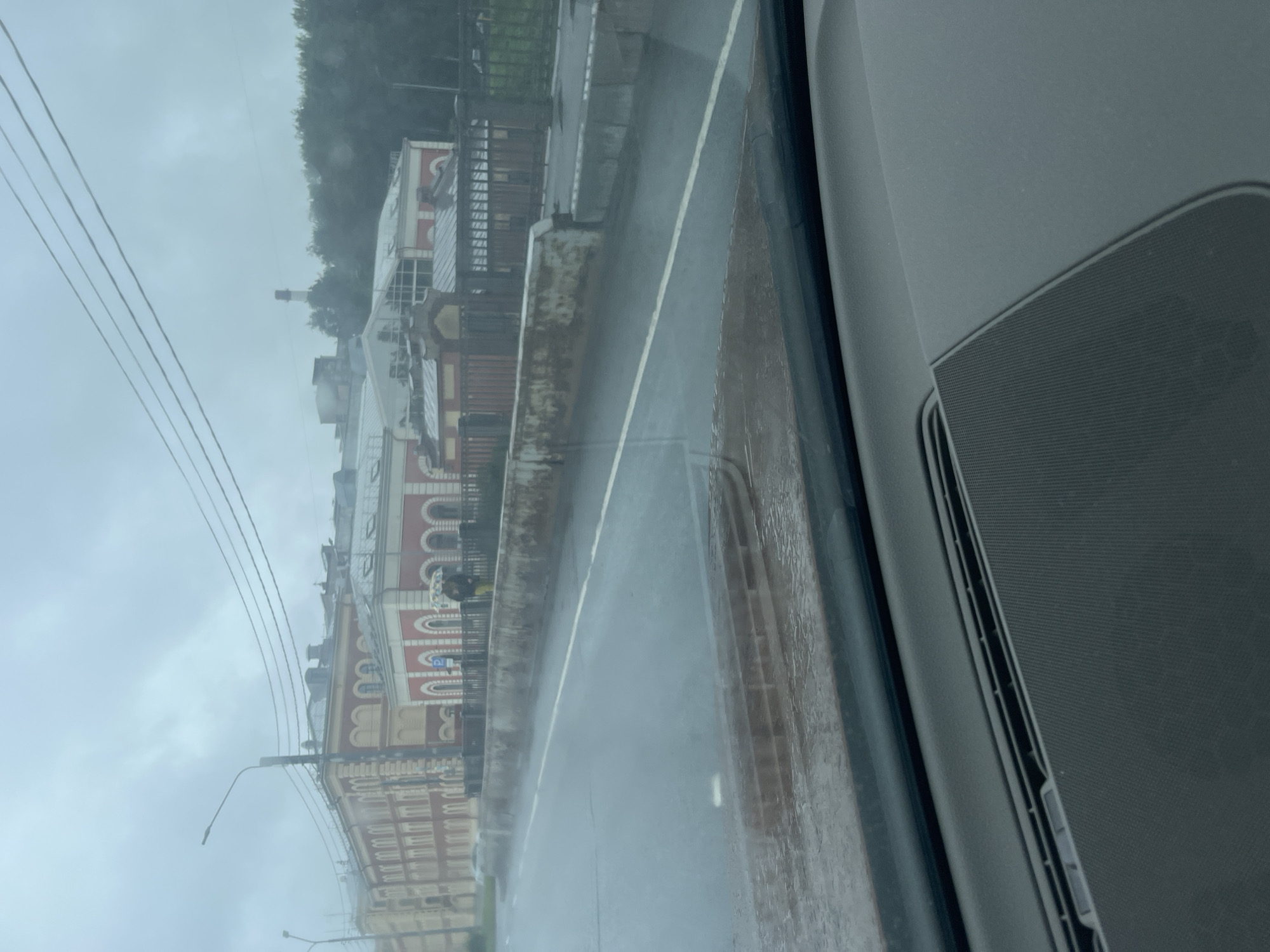 The image was taken from inside a vehicle, looking out at a cloudy day in Saint Petersburg, Russia. A wet road is visible in the foreground, with a white line marking a lane. Beyond the road, there is a stone retaining wall, and behind that, a sidewalk with a fence. Further in the background, a large, old building with red and yellow facades and arched windows is seen. To the right of this building, there are trees and a distant industrial chimney. Two people are walking on the sidewalk, one holding a colorful umbrella, indicating it is raining. The overall atmosphere is somber, consistent with overcast weather.