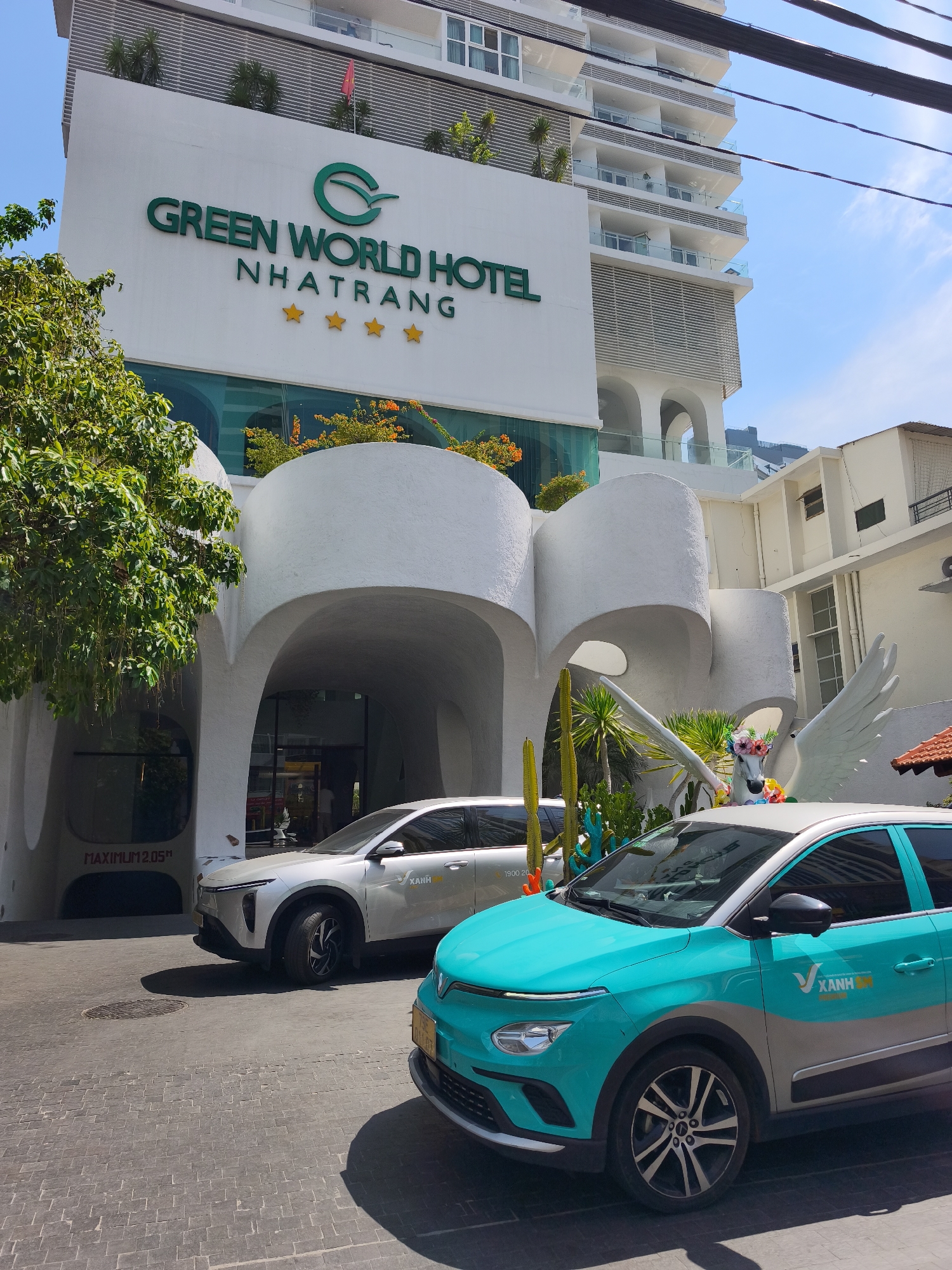 The image shows the exterior of the Green World Hotel Nha Trang, a modern white building with a distinctive curved entrance. The hotel's name is displayed in large green letters above the entrance. In front of the hotel, there are two cars parked on a paved street. One is a white SUV, and the other is a teal and silver car with "XANH SM" branding. A large white sculpture of a winged horse decorated with flowers stands near the entrance. Trees and plants, including cacti, are visible around the entrance and in front of the building. The sky is blue with some clouds, indicating a sunny day. The scene is likely a street view in Nha Trang, Vietnam.