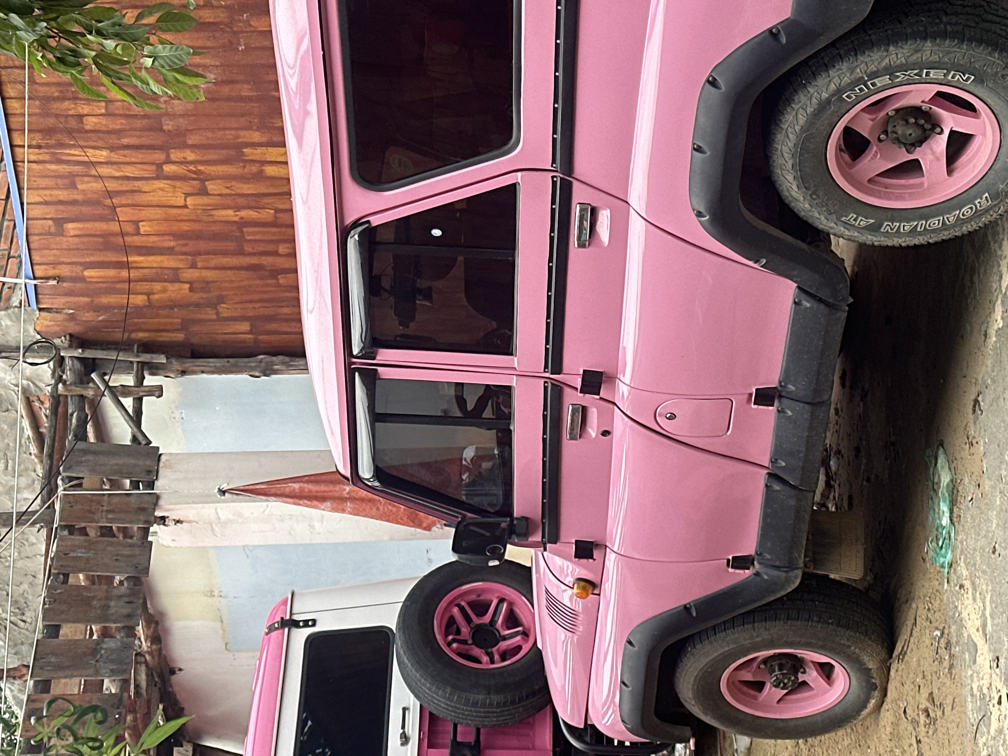 A pink Toyota Land Cruiser is parked on a street in Phu Thuy Ward, Vietnam. The vehicle has black fender flares and pink rims. The tires are Nexen Roadian ATs. The car is next to a white vehicle and partially obscured by a wooden structure, possibly scaffolding, and some tropical vegetation. In the background, there is a wall made of reddish-brown bricks. The ground is dirt.