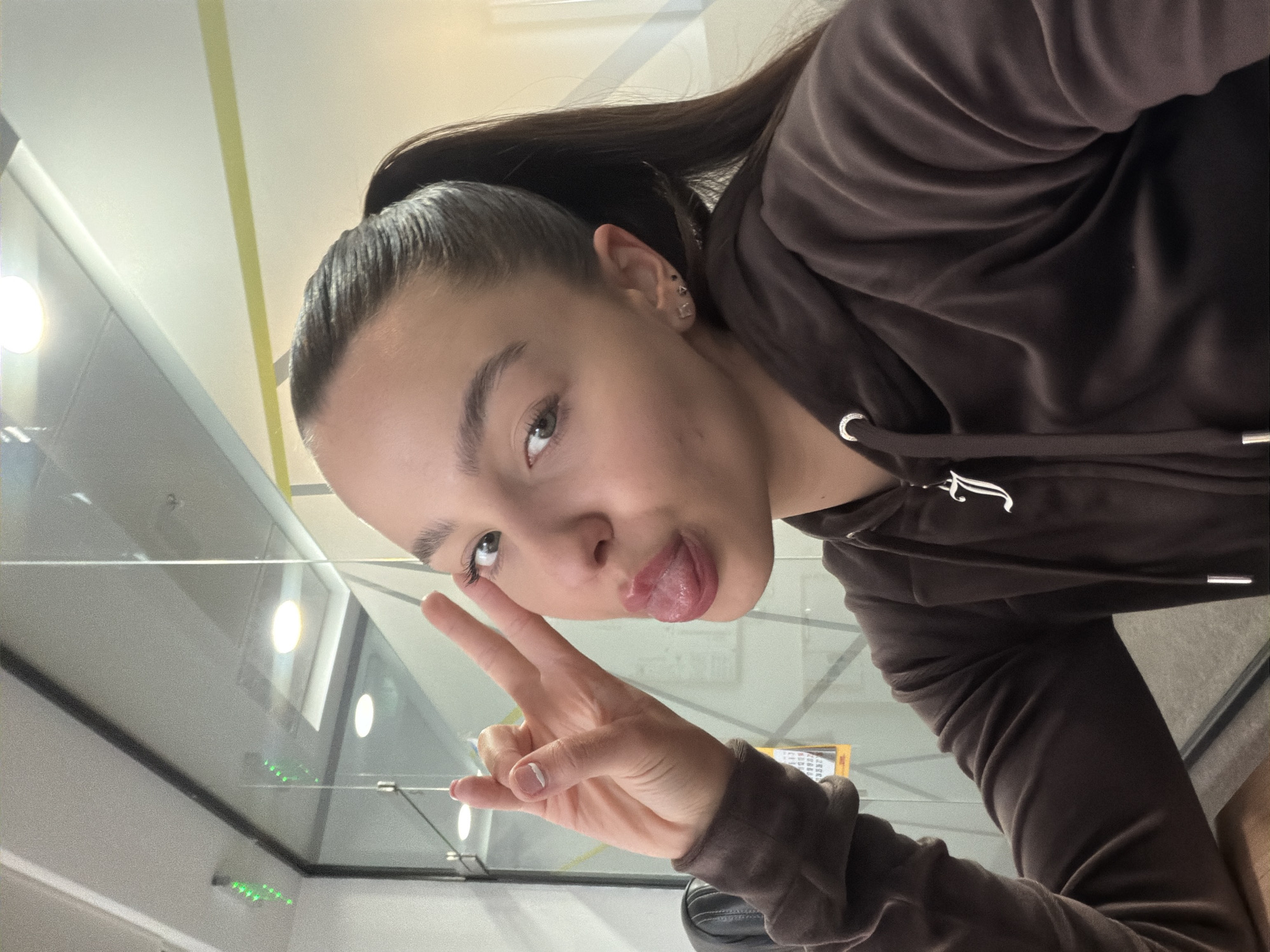A young woman with dark hair pulled into a high ponytail is making a peace sign with her left hand while sticking out her tongue. She is wearing a brown velvet hoodie with the drawstring visible. The image is taken from a low angle, and the background appears to be an office setting with glass partitions. It is not possible to determine if the location is Sofia, Bulgaria based on the provided image.
