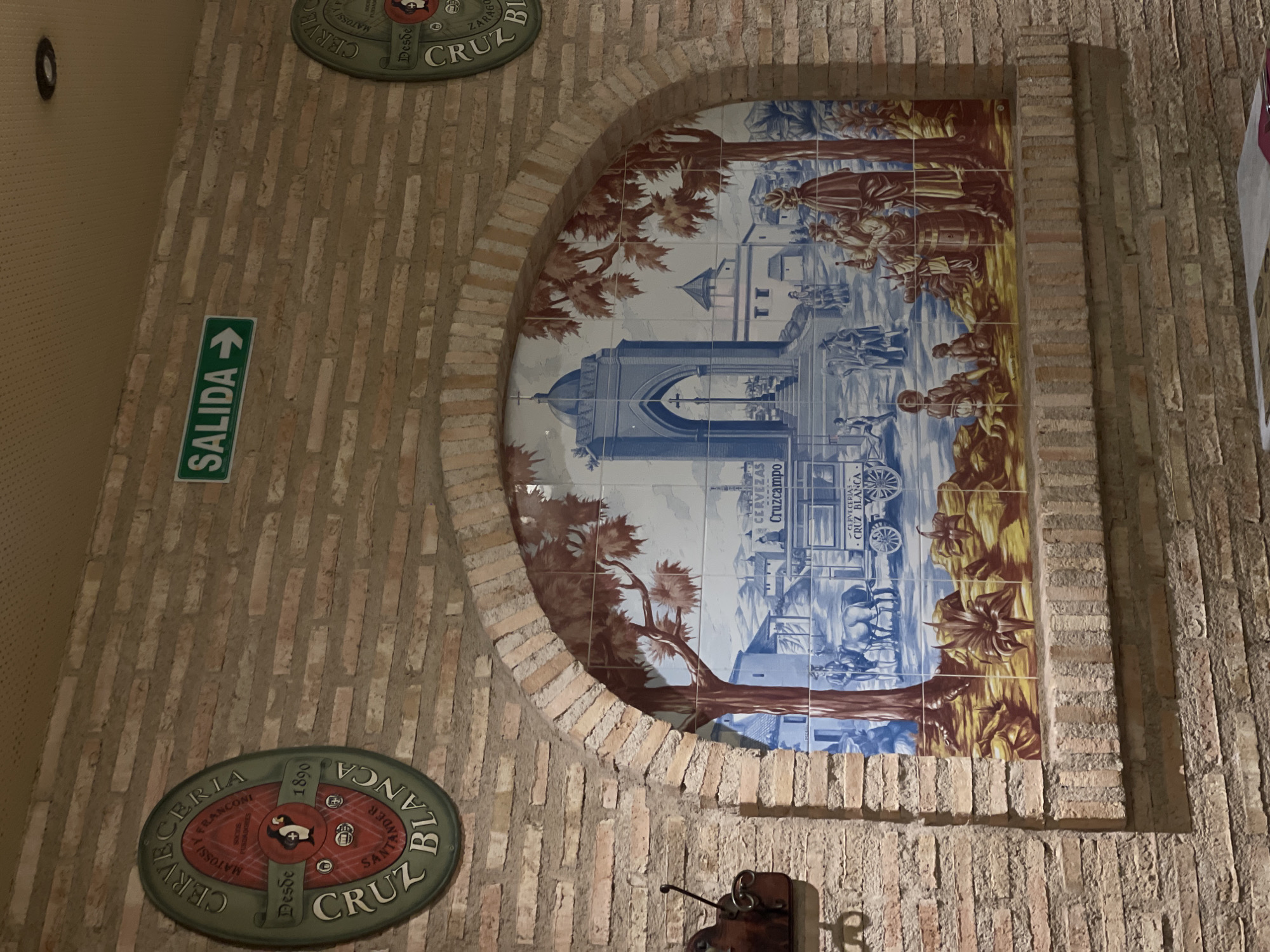 This is a medium shot, eye-level, interior photograph of a brick wall in Castelló De La Plana, Spain. The wall features a large, arched ceramic tile mural depicting a historical scene with buildings, trees, and people in traditional attire. A horse-drawn carriage is also present in the mural. Above the mural, there is an "SALIDA" (EXIT) sign with an arrow pointing to the right. To the left of the mural, a circular sign with a penguin logo and the text "CERVECERIA CRUZ BLANCA" and "DESDE 1890" is mounted on the wall. To the right of the mural, another circular sign is partially visible. The overall environment appears to be a restaurant or bar.