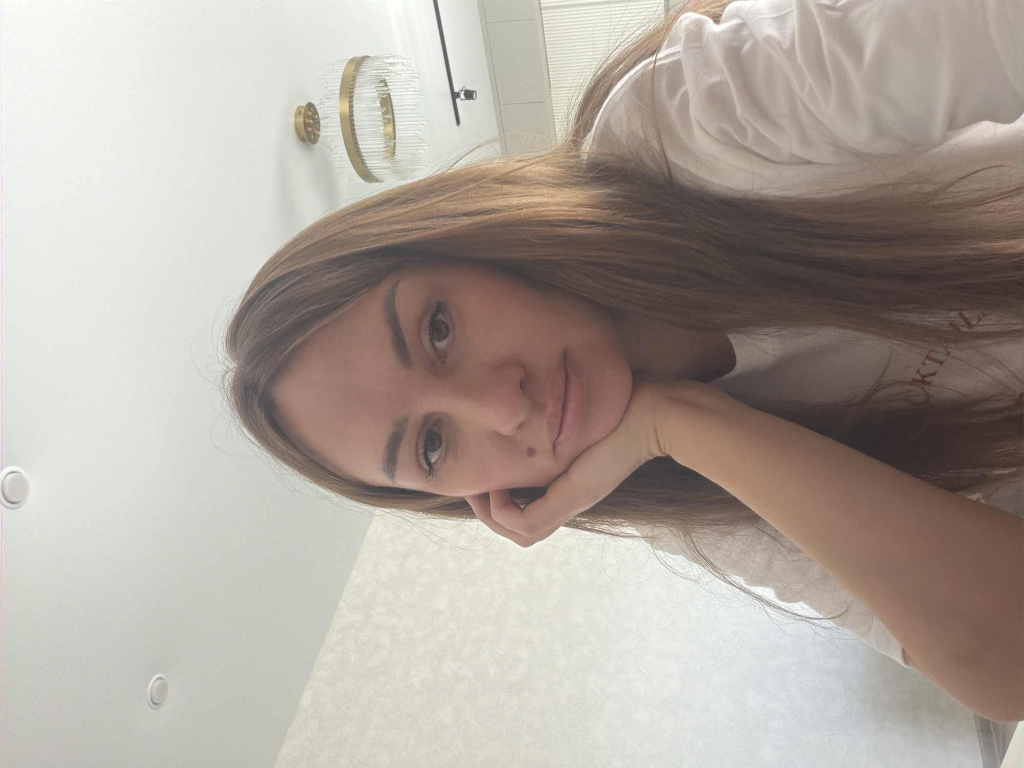 A woman with long brown hair is leaning her head on her hand, looking directly at the camera. She is wearing a white t-shirt. The background appears to be a well-lit room with a textured wall and a modern chandelier. There is also a glimpse of cabinetry and a ceiling light fixture. The scene is set indoors, likely in a residential environment. It is not possible to determine the specific city of Malinovka, Russia from the image alone.