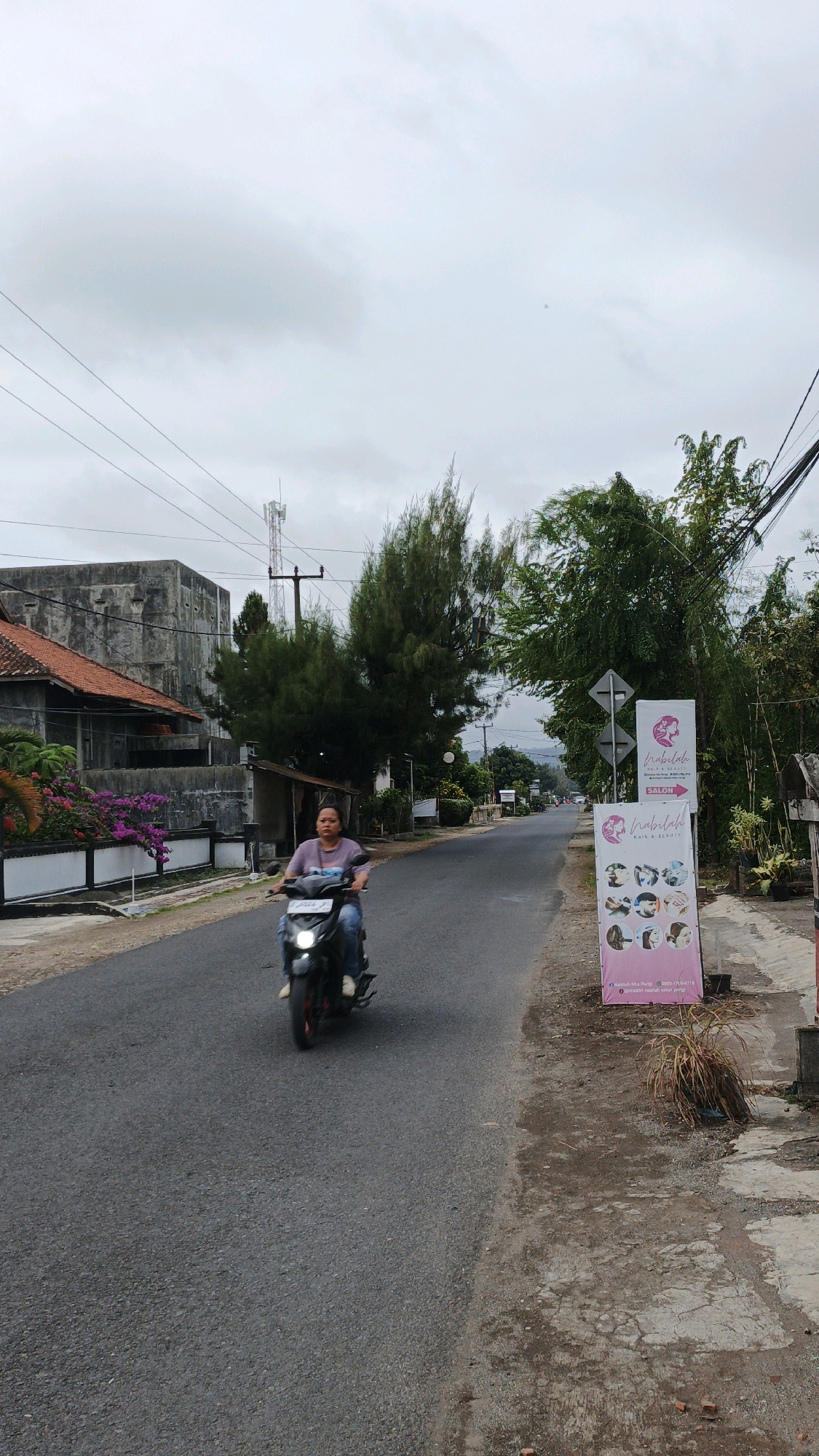 A woman rides a scooter down a paved road in Parigi, Indonesia. The road is lined with trees and buildings on both sides. A cell tower is visible in the background. A banner advertising a salon stands on the right side of the road. The sky is overcast. The woman is wearing a purple shirt and blue jeans.