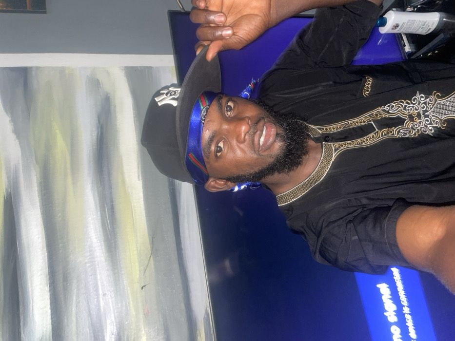 A Black man with a beard poses in a black shirt with gold accents and a black baseball cap with a blue patterned band. He appears to be indoors, standing in front of a television screen displaying the message "no signal, device is connected." A partially visible abstract painting is behind him on the wall. A bottle is visible in the upper right corner. The photo is tilted to the left.