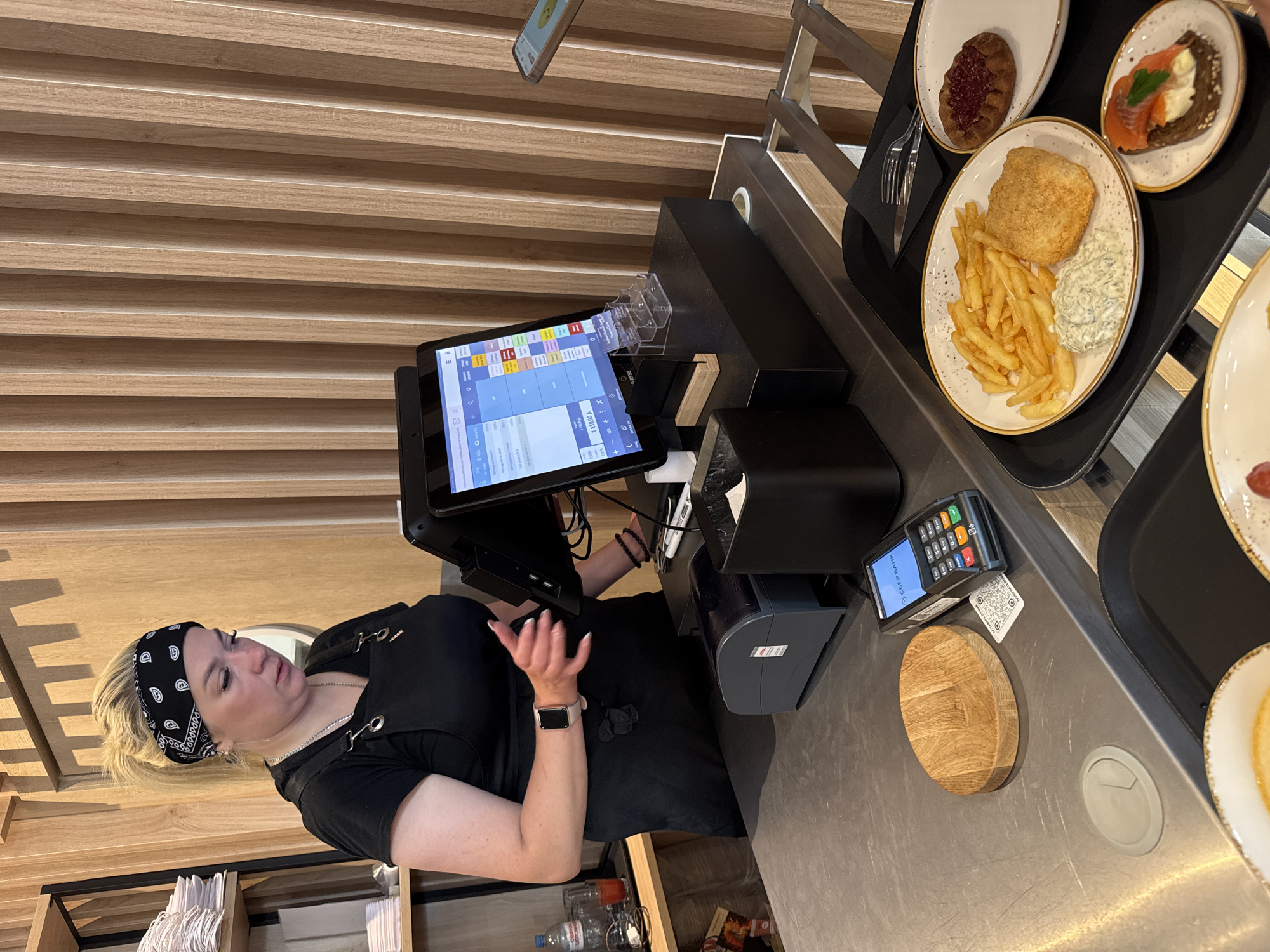 An overhead medium shot shows a female cashier with blonde hair pulled back and wearing a black bandana and apron, standing behind a counter in what appears to be a food establishment. She is looking down at a tablet computer that displays a sales interface. In front of her is a point-of-sale system, a credit card reader, and a wooden coaster. To the right, a tray holds a meal consisting of french fries, fried fish, and a small dish of sauce, along with a pastry and a piece of open-faced sandwich with salmon. The background features a wall of vertical wooden slats. The scene is set in Yakkima, Russia.